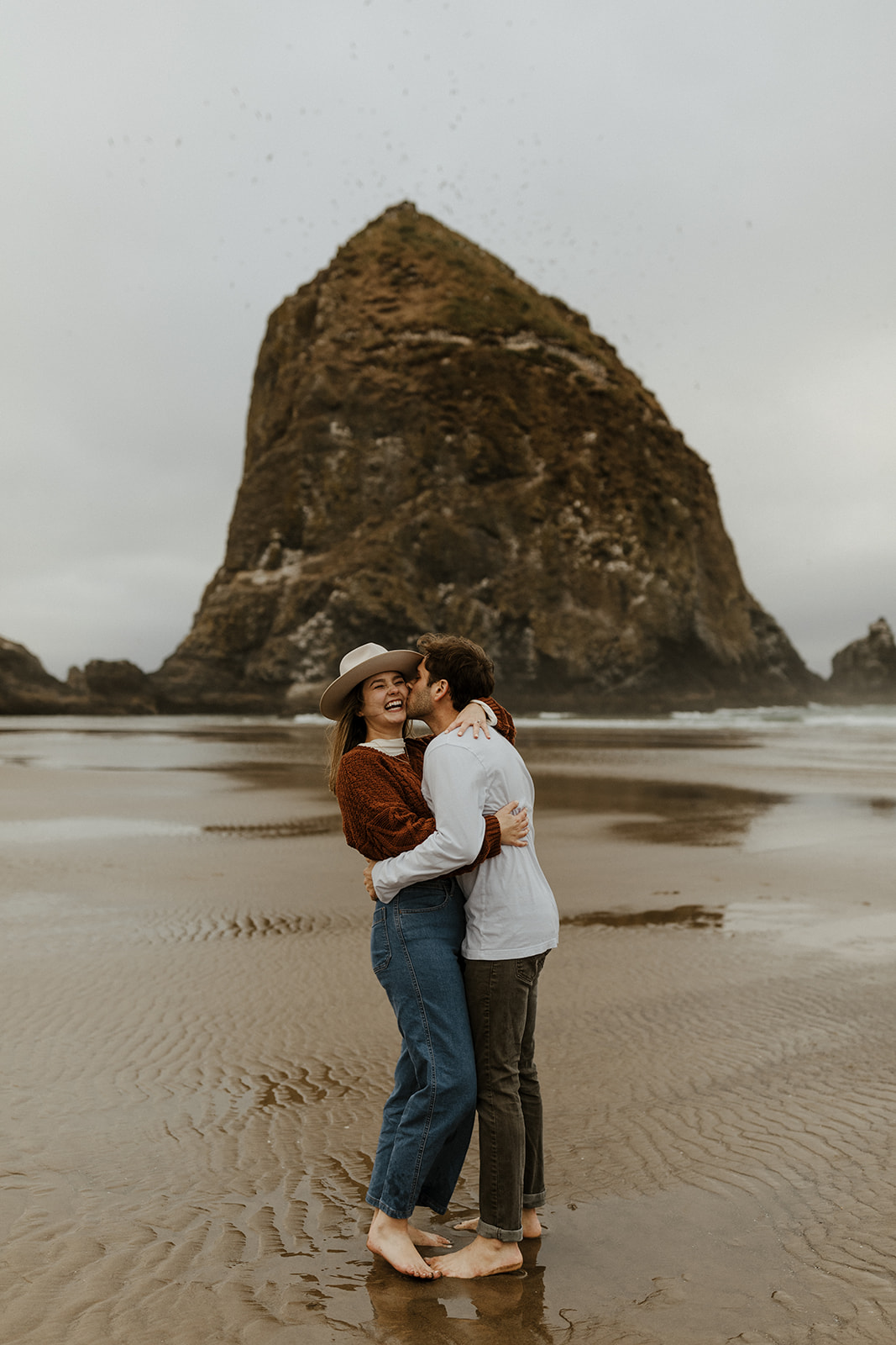 couple laughing at kissing on the beach with haystack rock in background