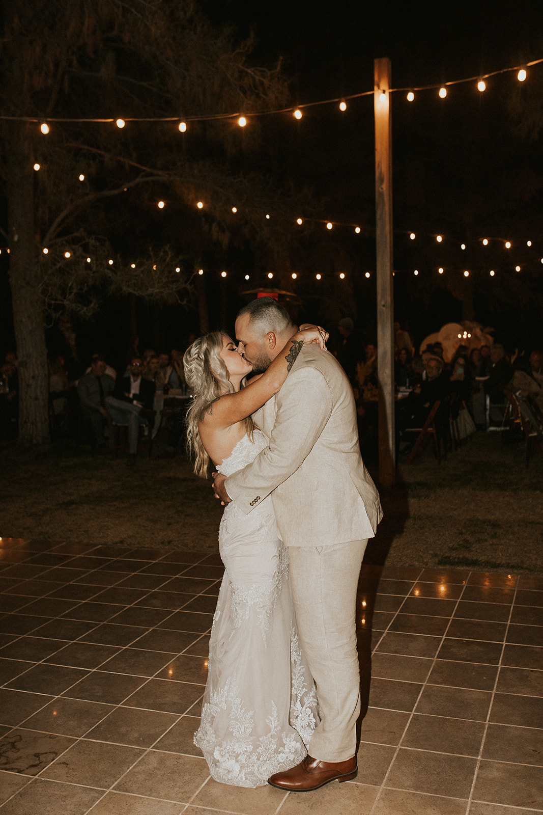 bridal couple kissing on dance floor with string lights above them 