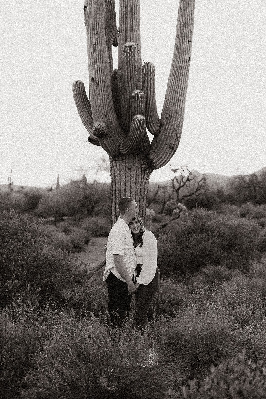 black and white of engaged couple hugging in front of giant cactus at lost dutchman state park
