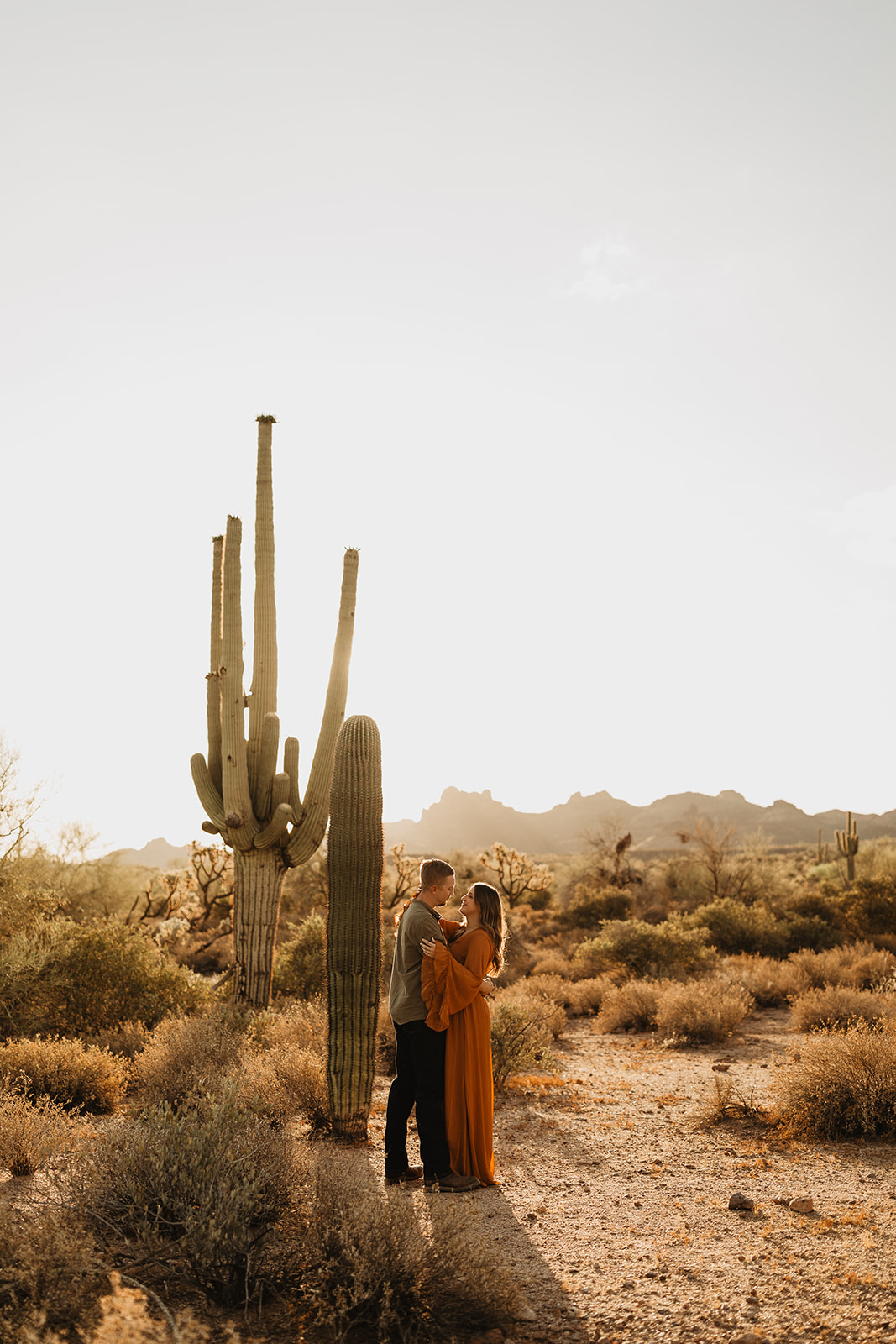 engaged couple holding each other in front of a cactus as the sun is rising 
