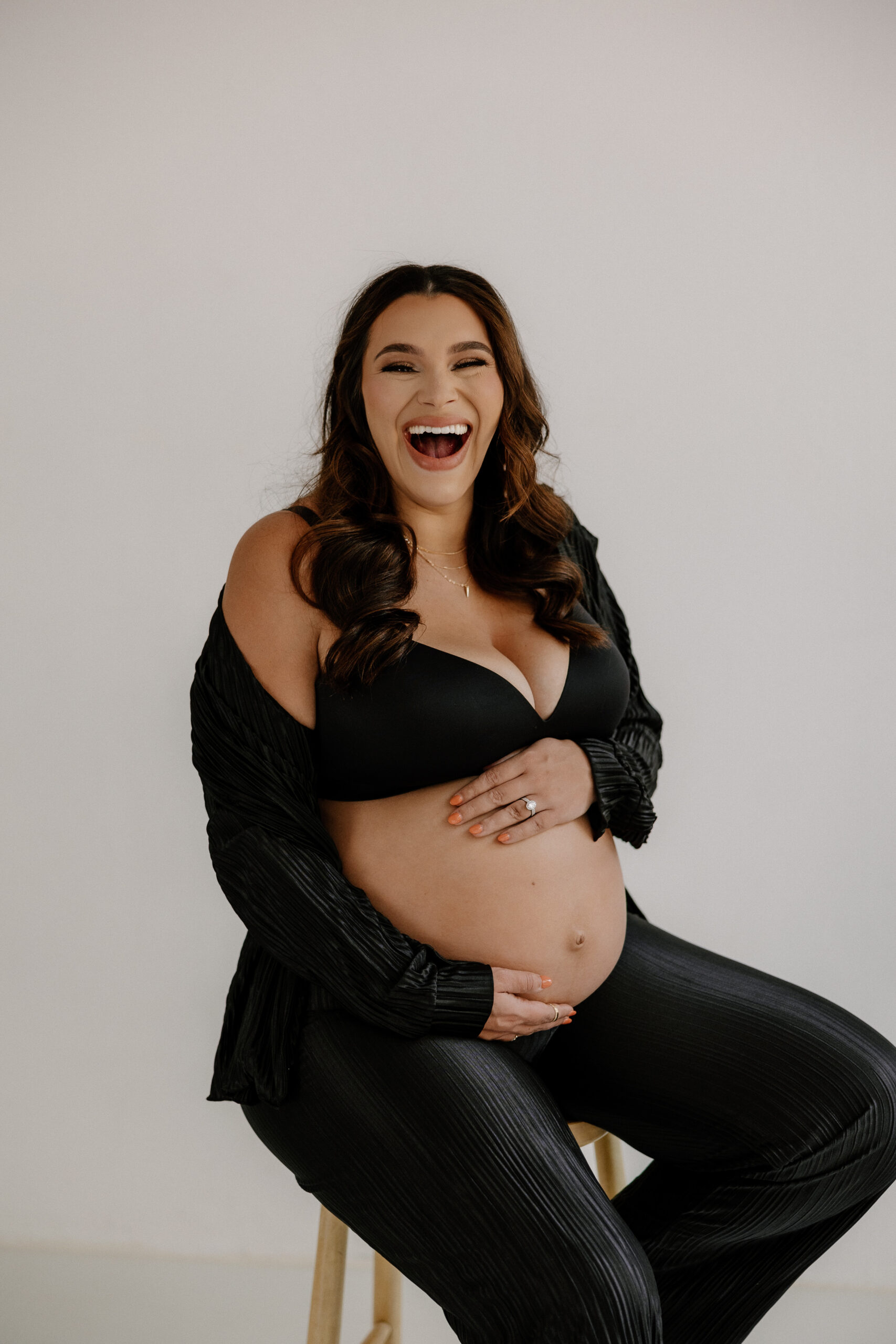 pregnant woman sitting on a stool holding her belly and laughing during editorial maternity photoshoot