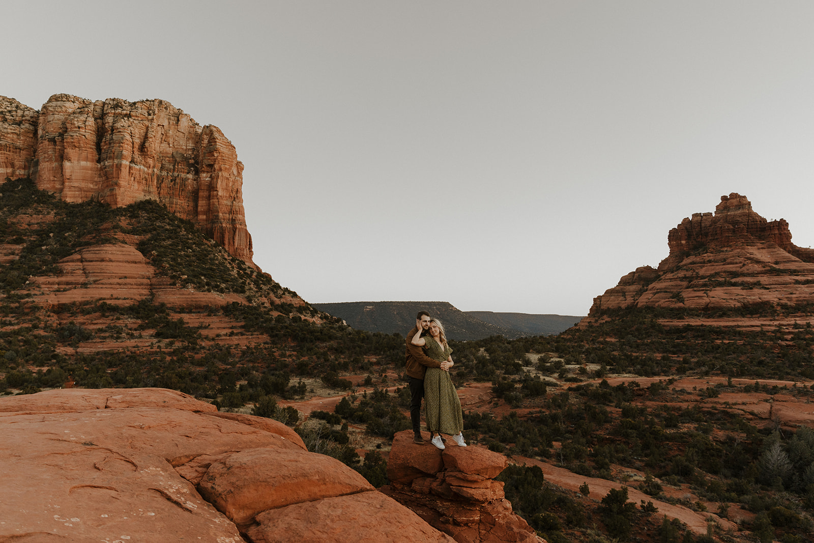 engaged couple holding each other during their sedona engagement photos with the red rocks and green plants behind the, 