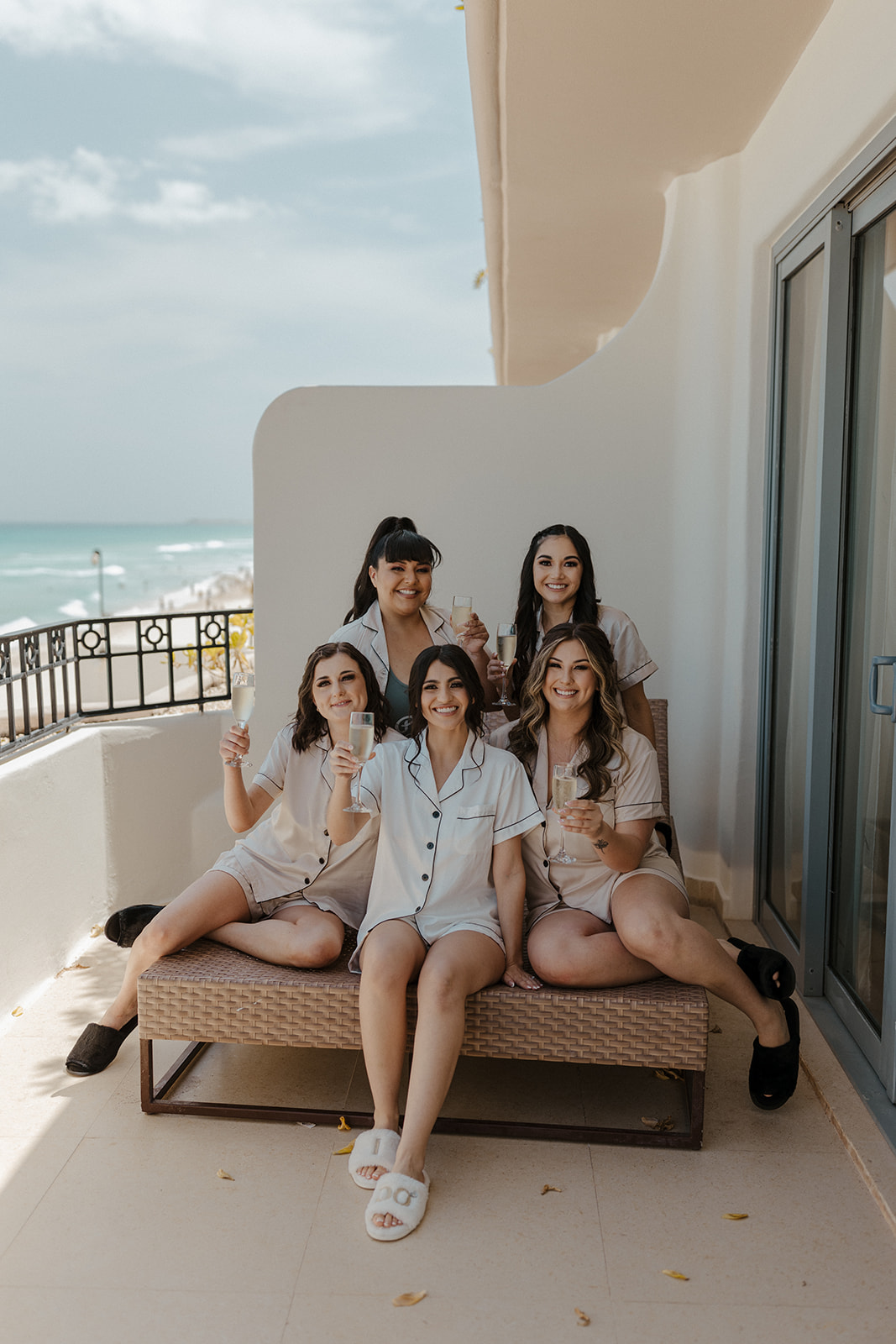 bride in white silk pajamas poses with bridesmaids in chrome silk pajamas on the patio overlooking the ocean