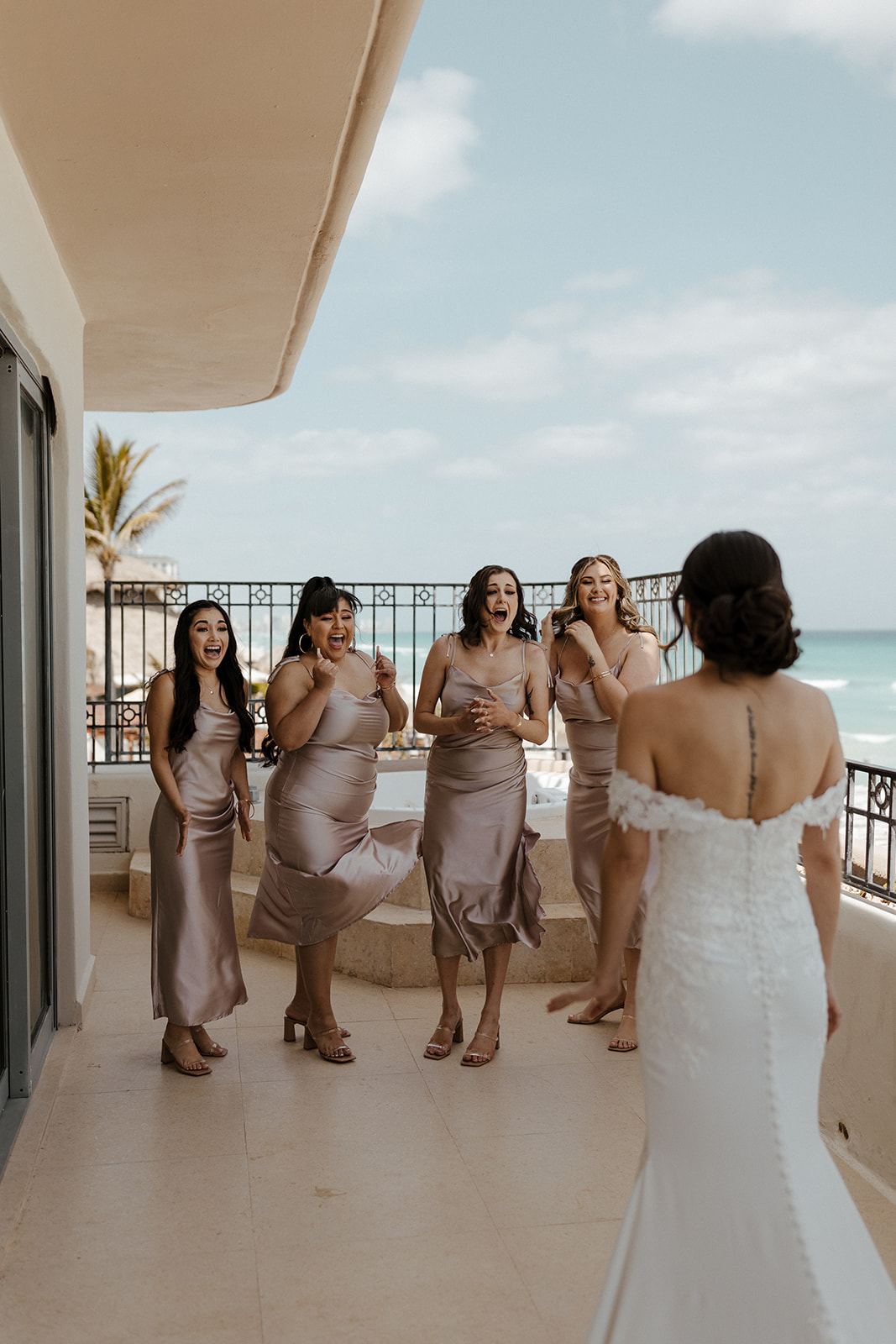 bride in lacy white dress poses for first look with bridesmaids in chrome dresses on the patio overlooking the ocean