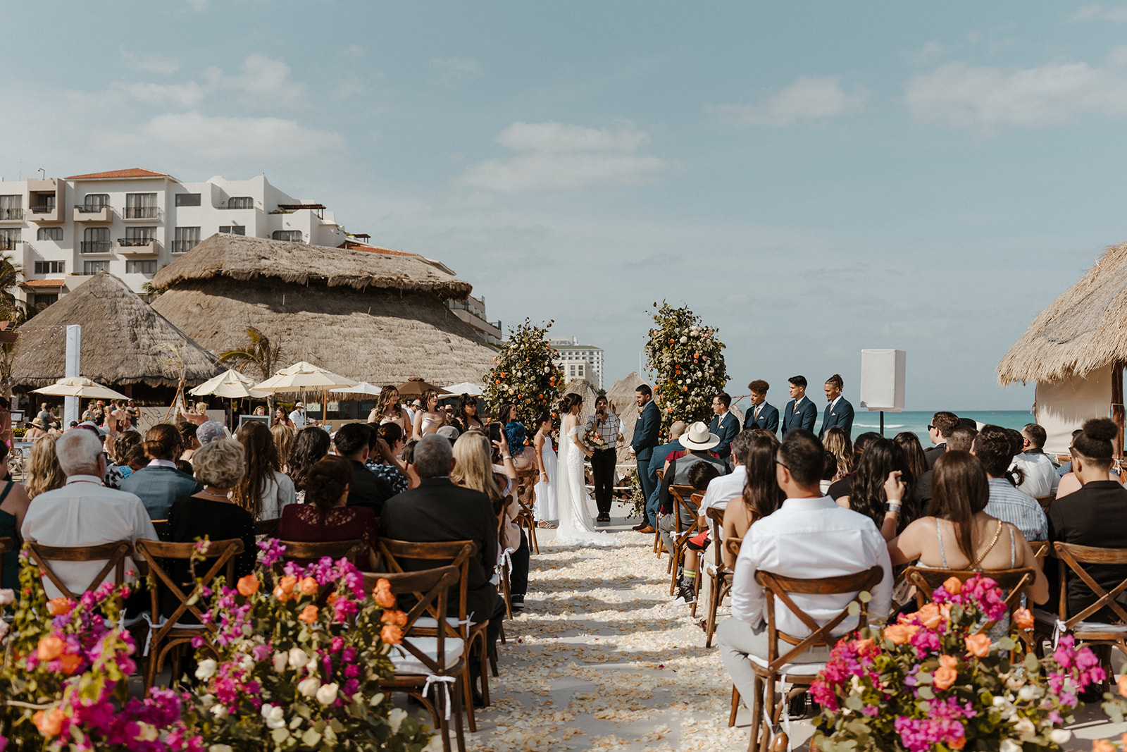 bride and groom pose during their destination wedding in cabo surrounded by pink, orange, and white flowers and their loved ones