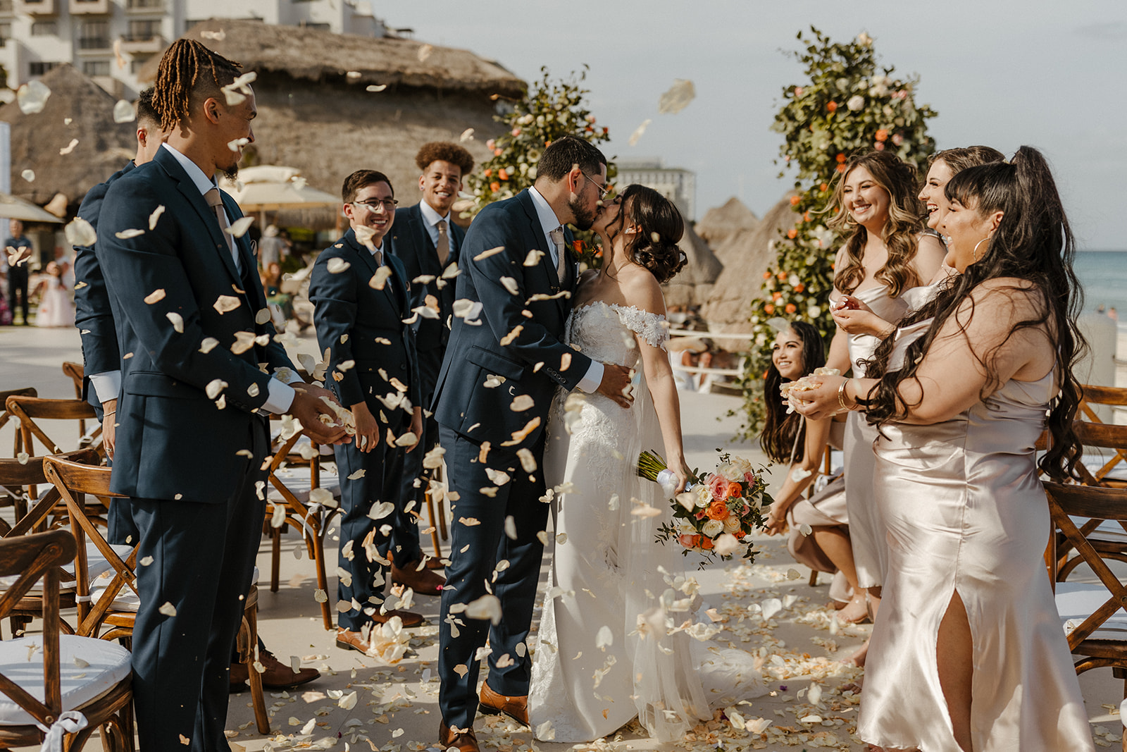 bride and groom pose during their destination wedding in cabo surrounded by pink, orange, and white flowers and their loved ones