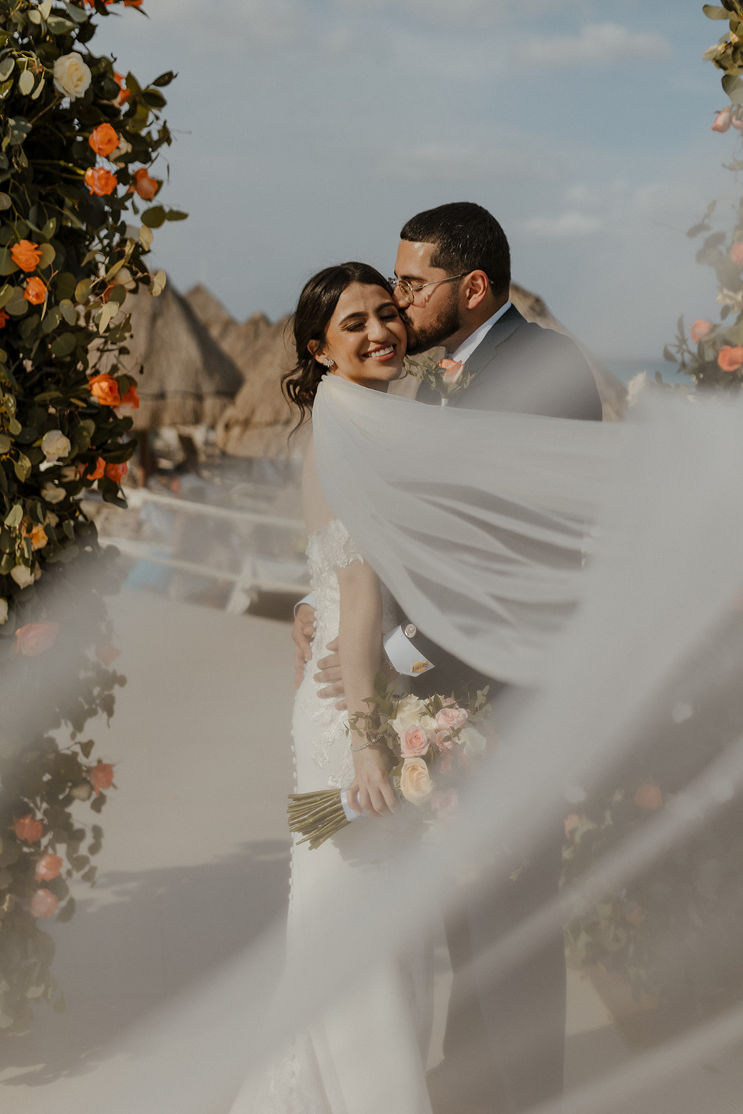 bride and groom pose during their destination wedding in cabo surrounded by pink, orange, and white flowers and their loved ones