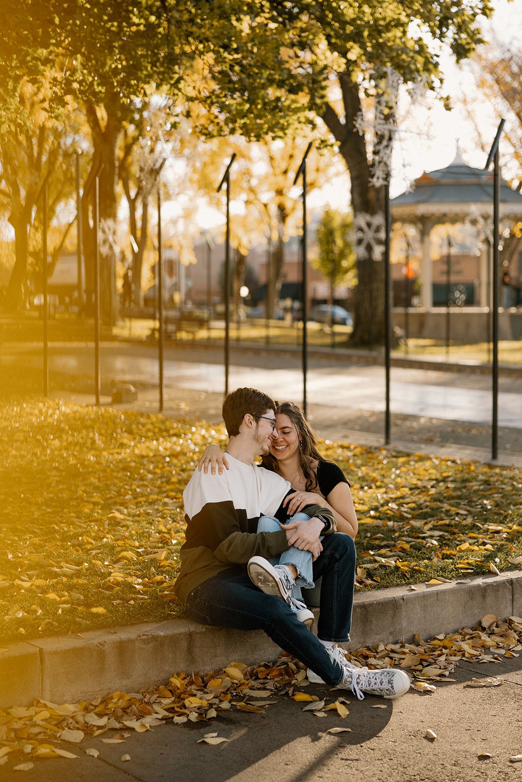 couple sits on sidewalk holding each other during the fall season with orange and yellow leaves on the ground