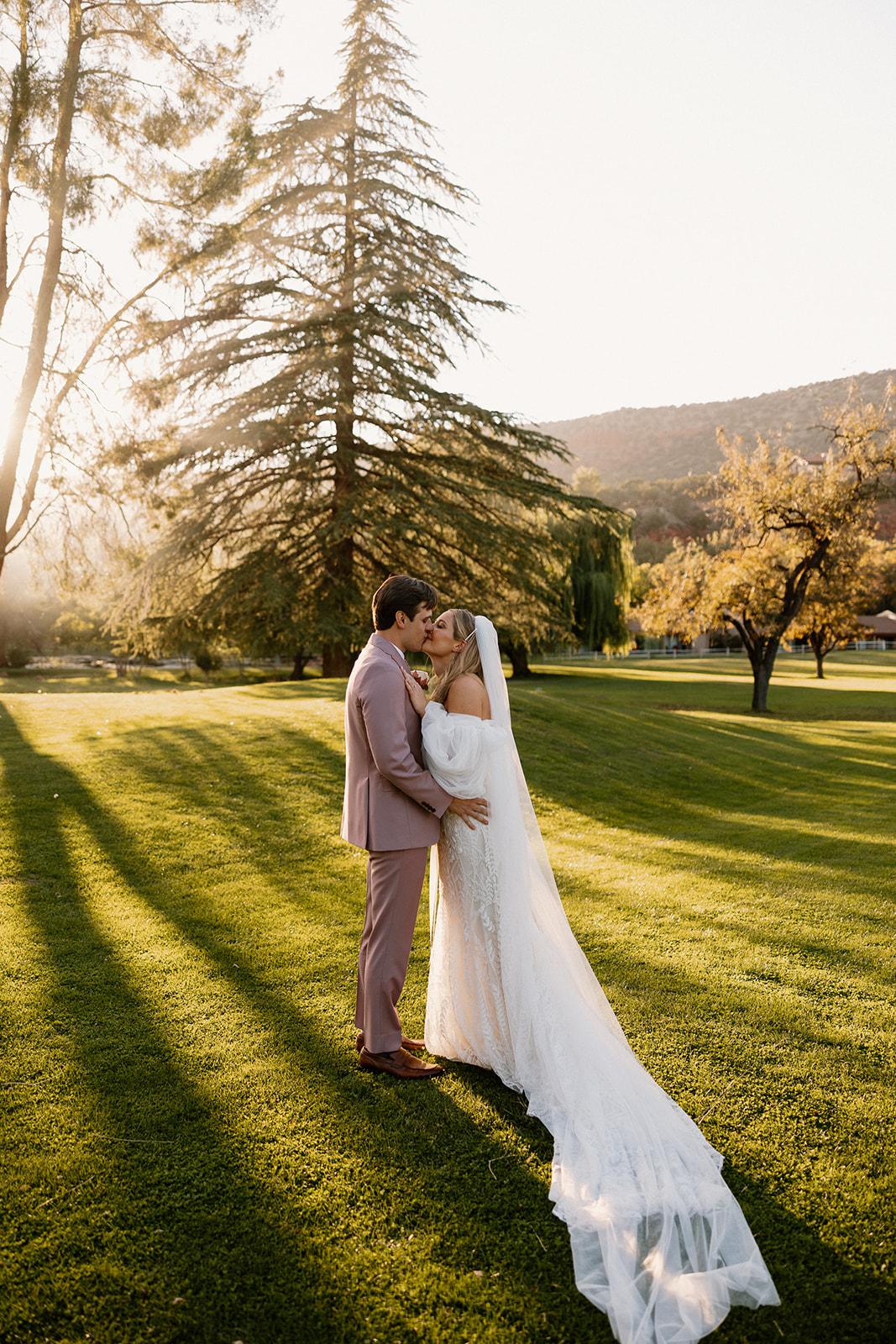 Happy couple kiss while standing in grassy area while wearing wedding attire