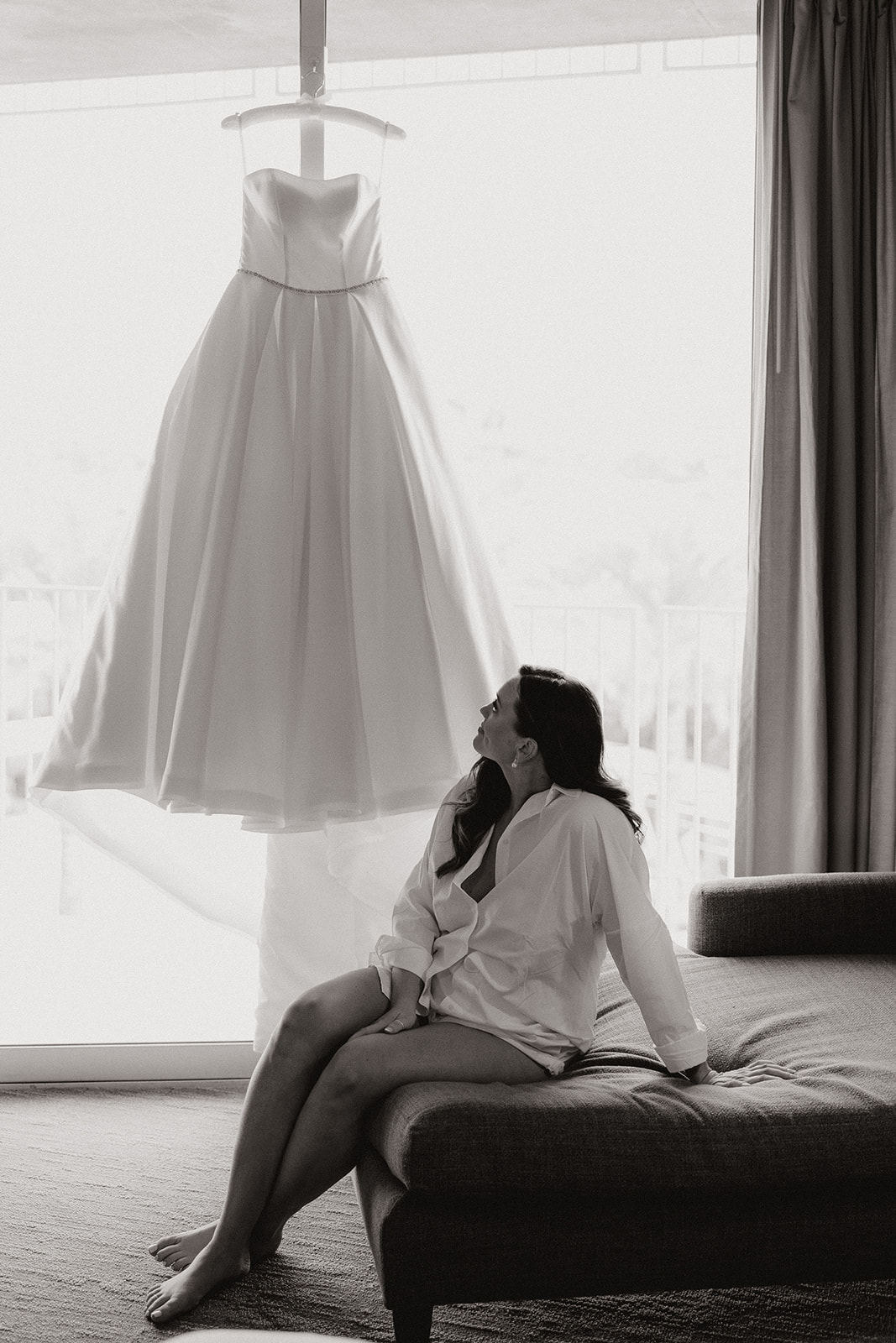 bride sitting on couch looking up at wedding dress hanging in front of window