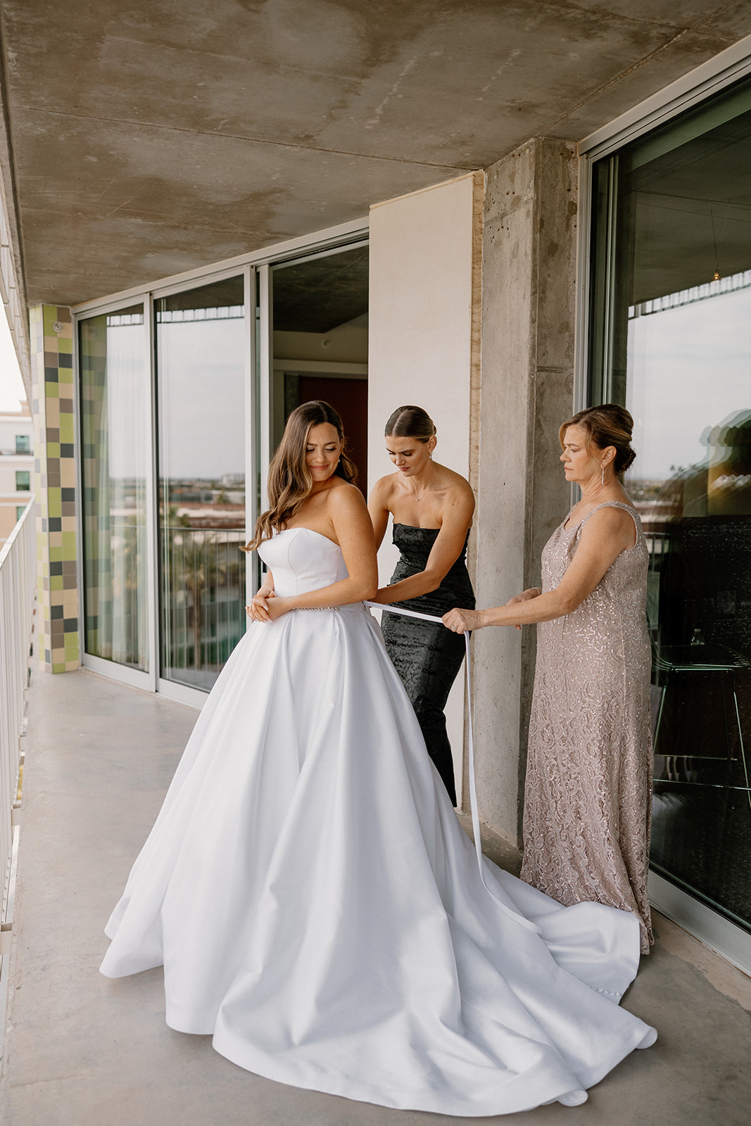 bride getting into strapless bridal gown with mother and bridesmaid helping her tie the back 