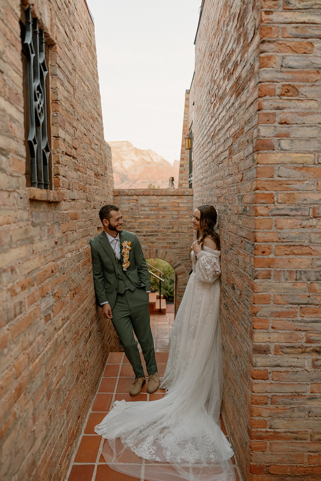 bride leans against brick wall looking at groom while groom leans against opposite brick wall looking at bride