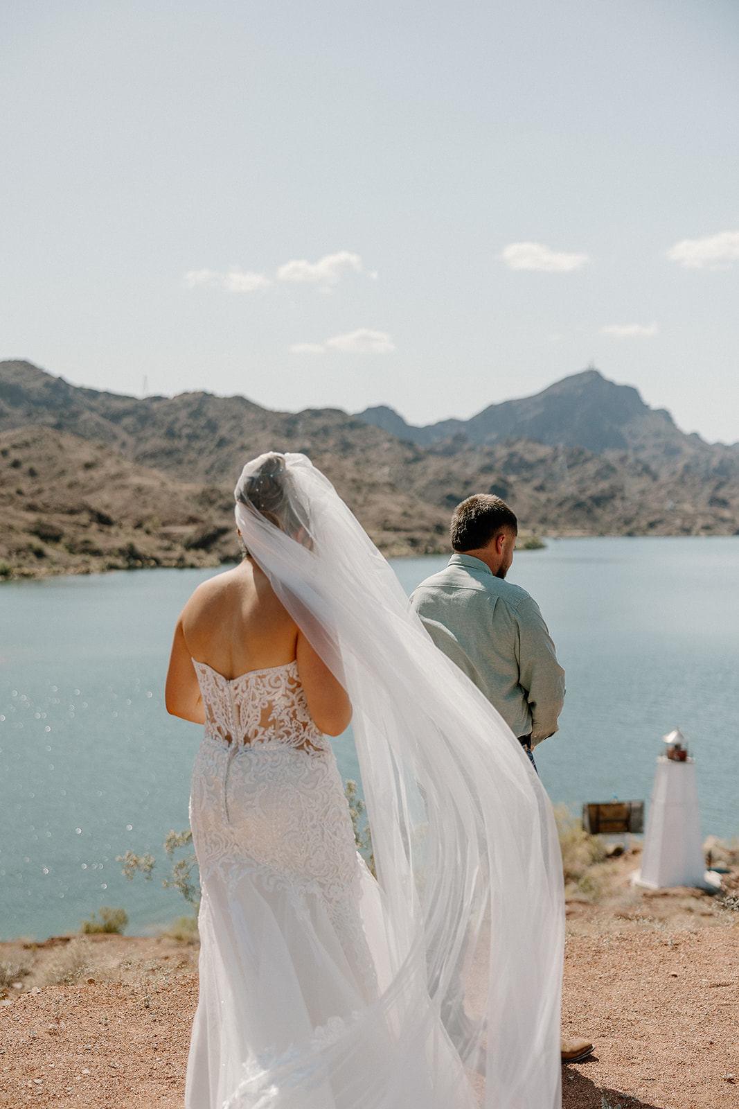 A bride walks behind her groom along a cliffside path, her veil trailing dramatically in the wind above a scenic Arizona lake.