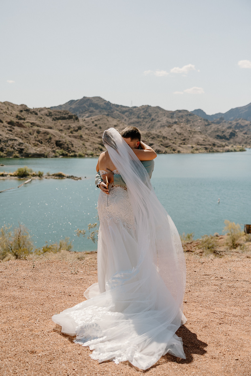 A newlywed couple stands arm-in-arm overlooking a bright blue lake, soaking in their just-married moment.