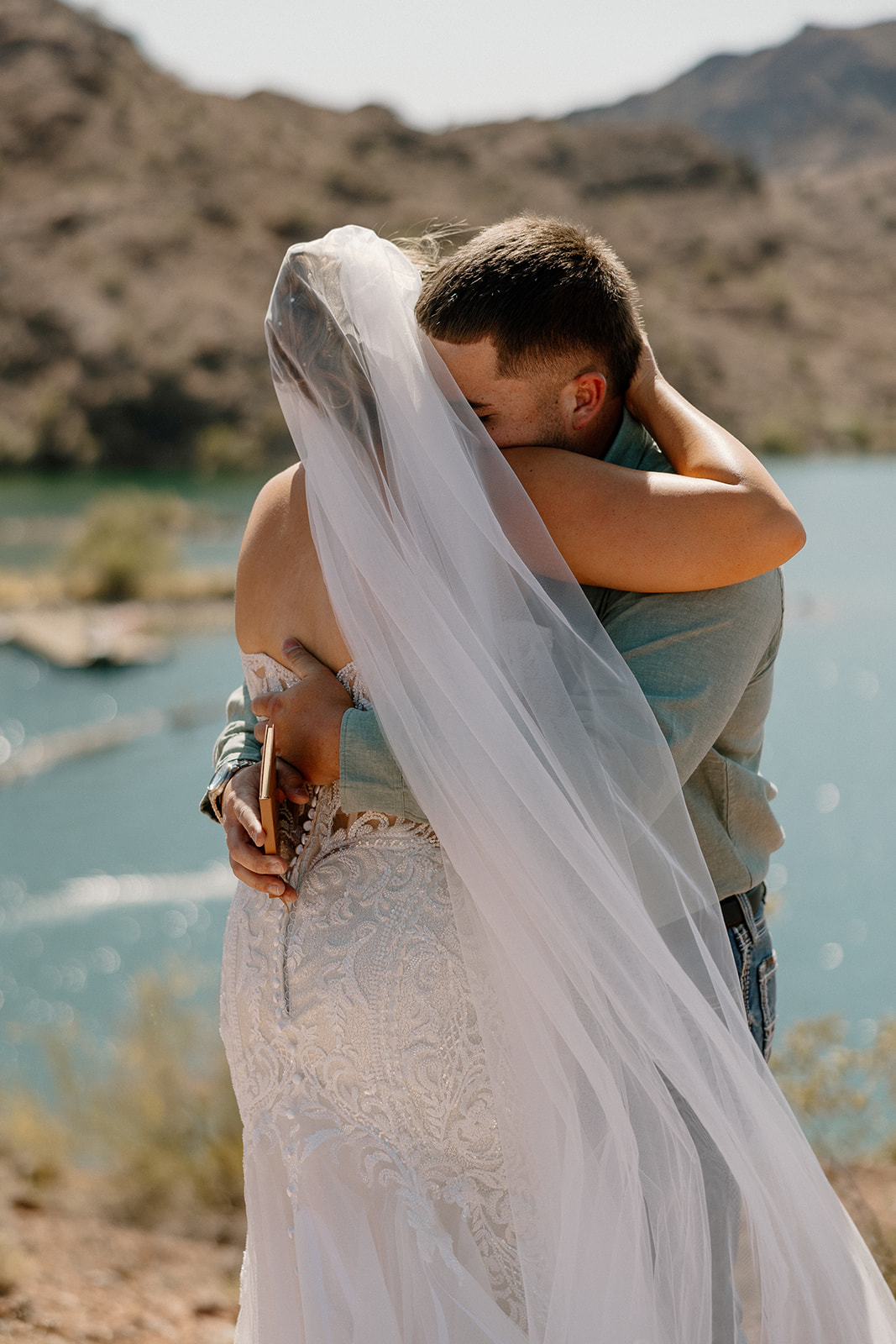 Close-up of a bride and groom hugging near the water, the bride clutching her vows while her veil flows in the breeze.