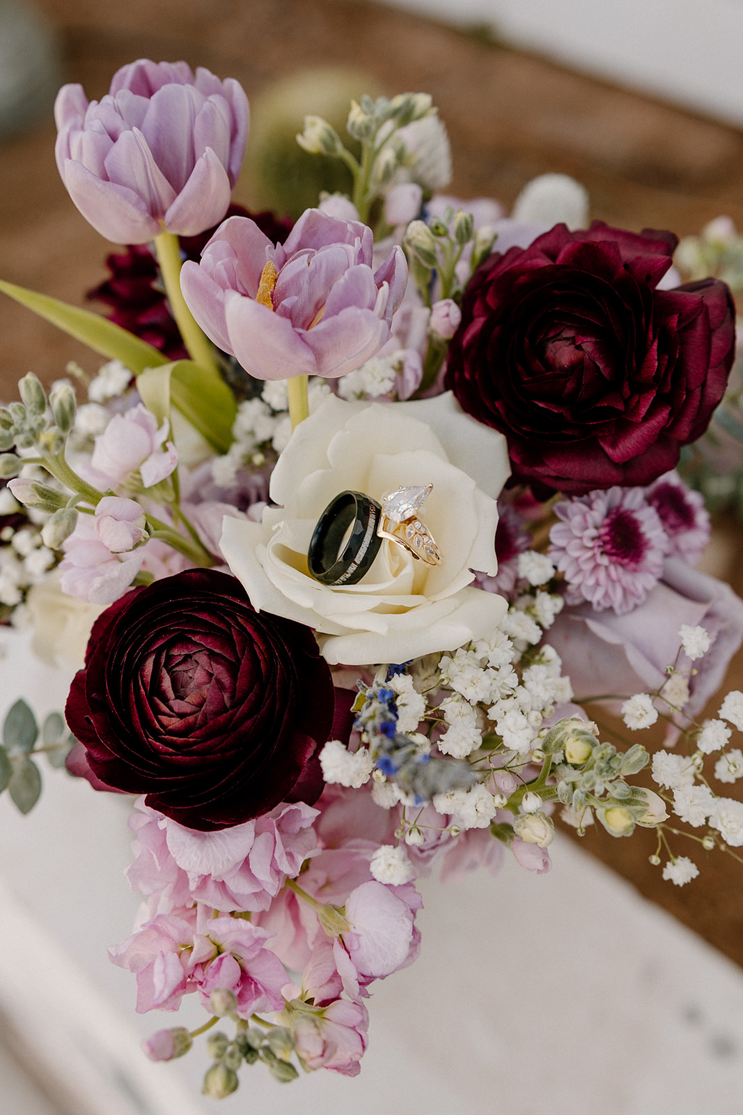 Wedding rings placed on white rose in a bridal bouquet with burgundy and lavender flowers at The Cove at Bartlett Lake wedding.