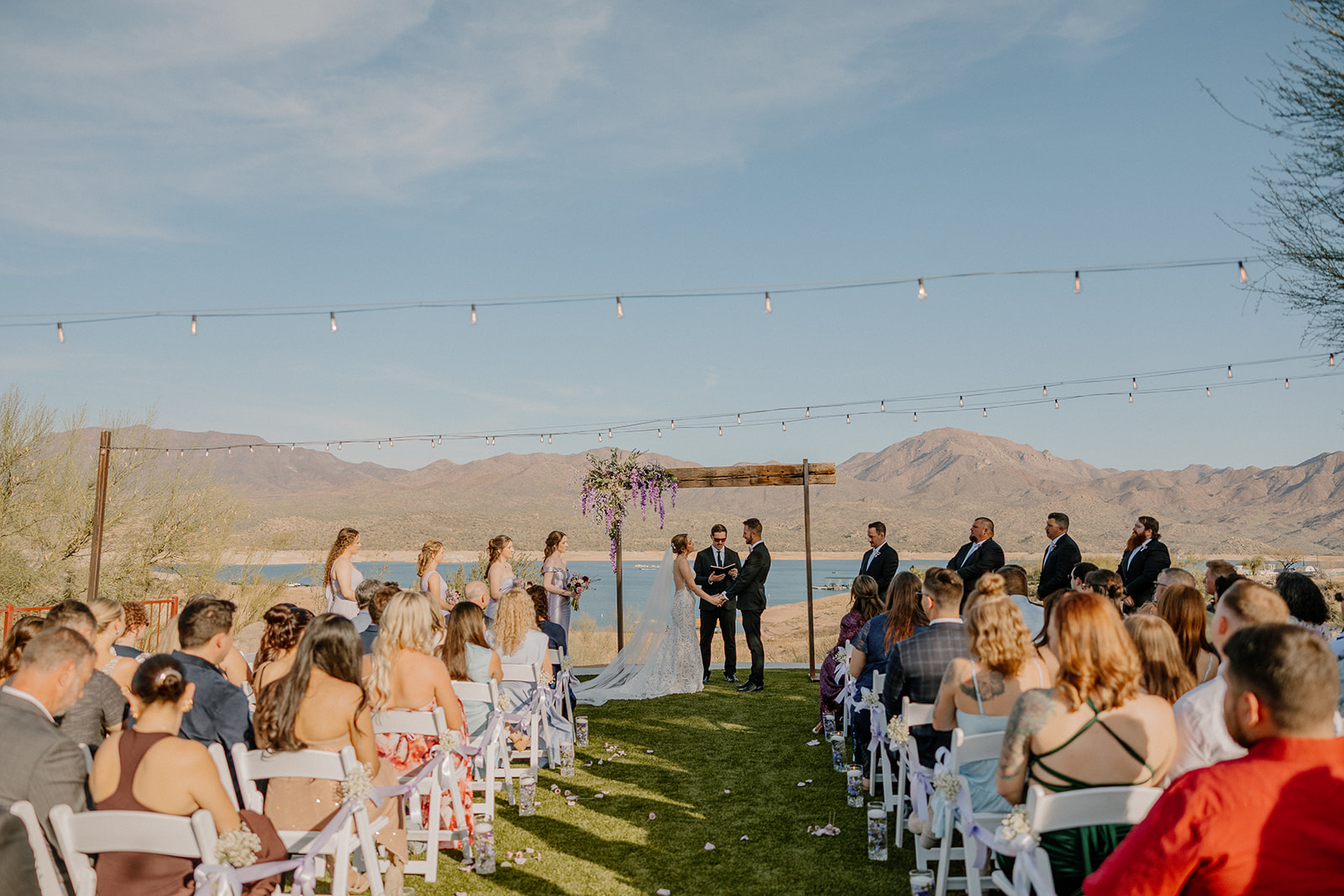 Wide shot of a wedding ceremony overlooking the water at The Cove at Bartlett Lake with string lights and mountain views.