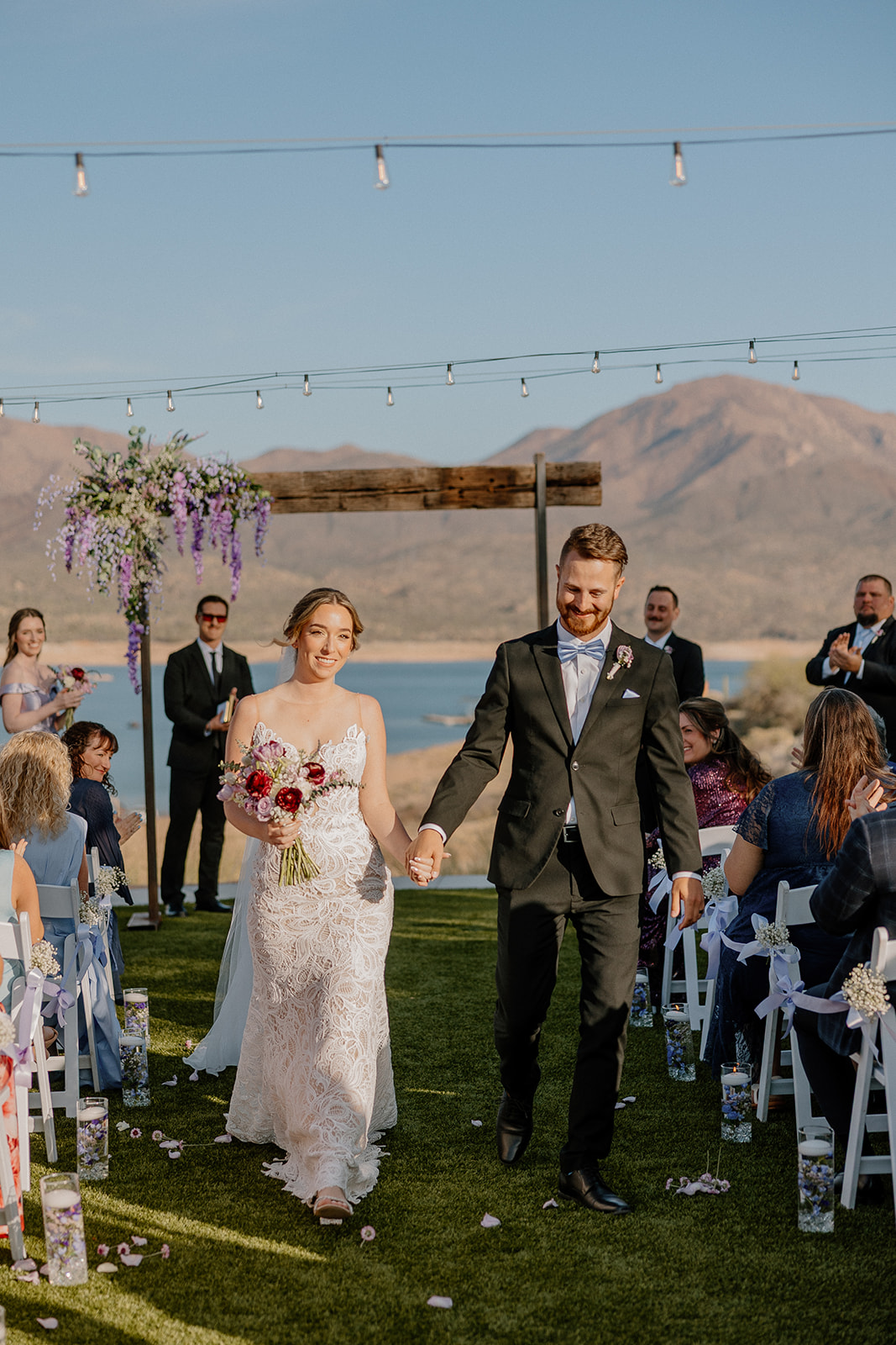 A joyful couple exits their ceremony, hand in hand, in front of a lake and mountain backdrop—surrounded by guests on a lush green lawn.