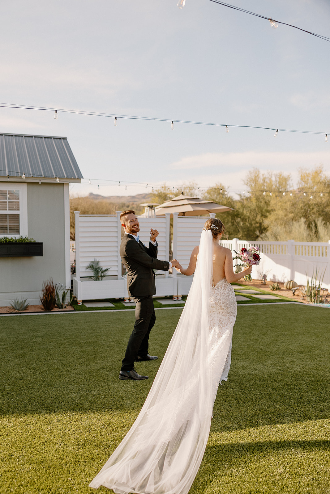 A playful moment between a bride and groom on a modern lawn, twirling under bistro lights—sunny skies and good vibes.
