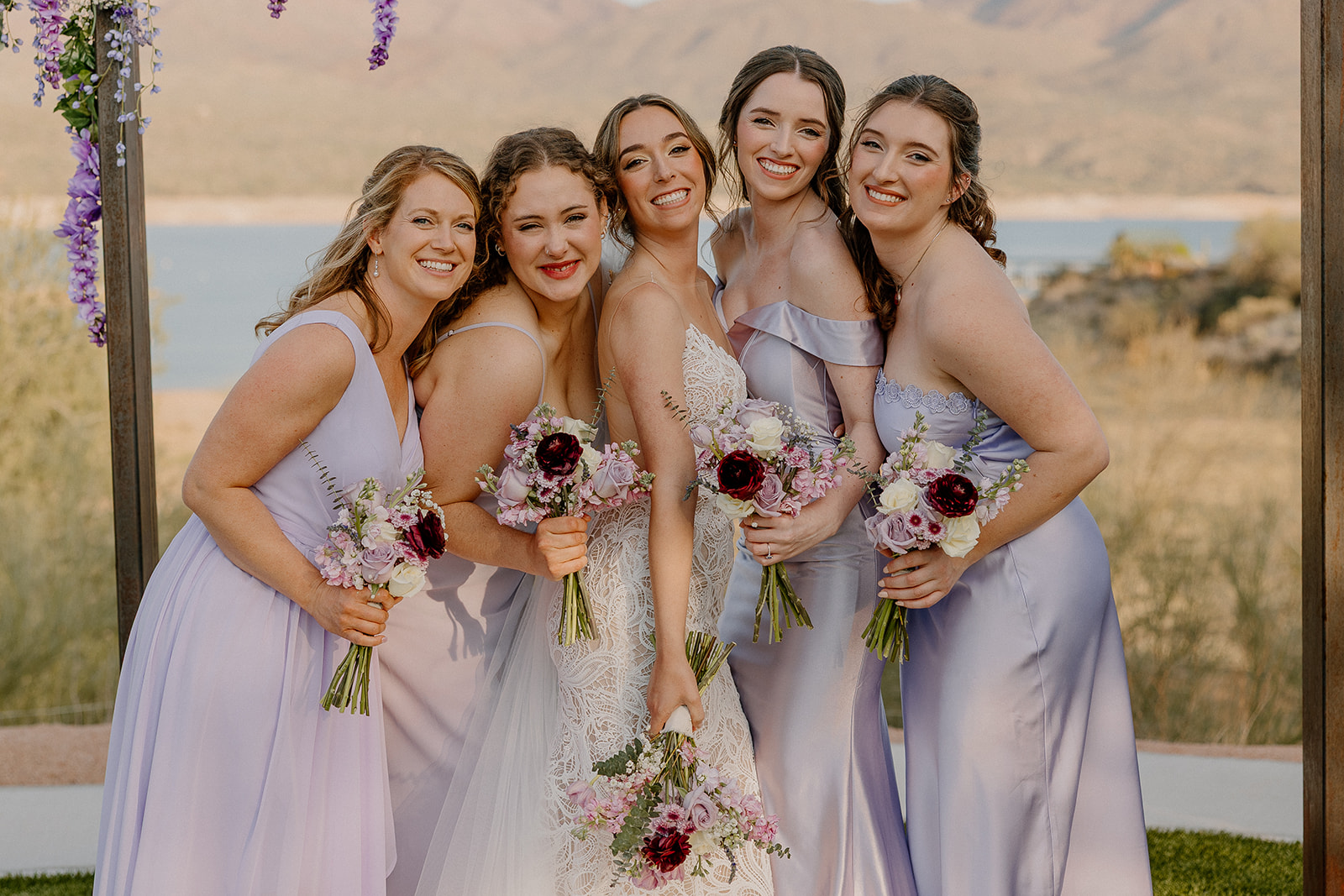 Bride and bridesmaids in lavender dresses holding pastel bouquets, posing at The Cove at Bartlett Lake wedding with a scenic desert lake backdrop.