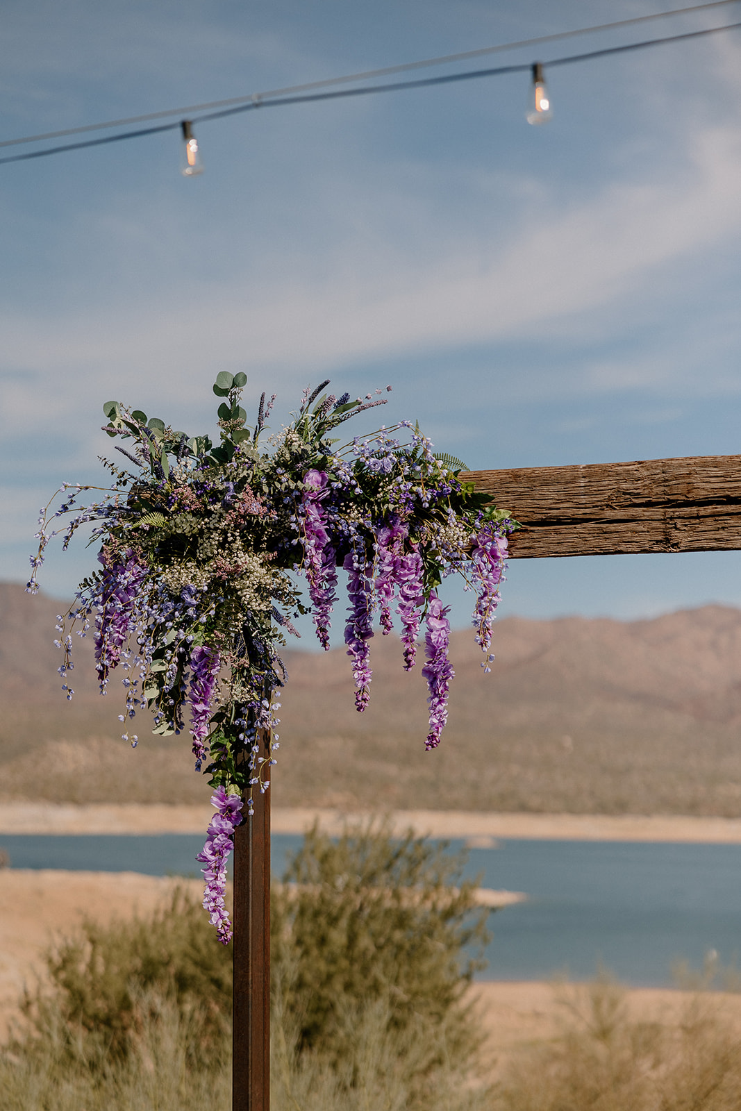 A rustic wooden ceremony arch adorned with cascading purple florals, with the lake and Arizona mountains stretching into the horizon behind it.
