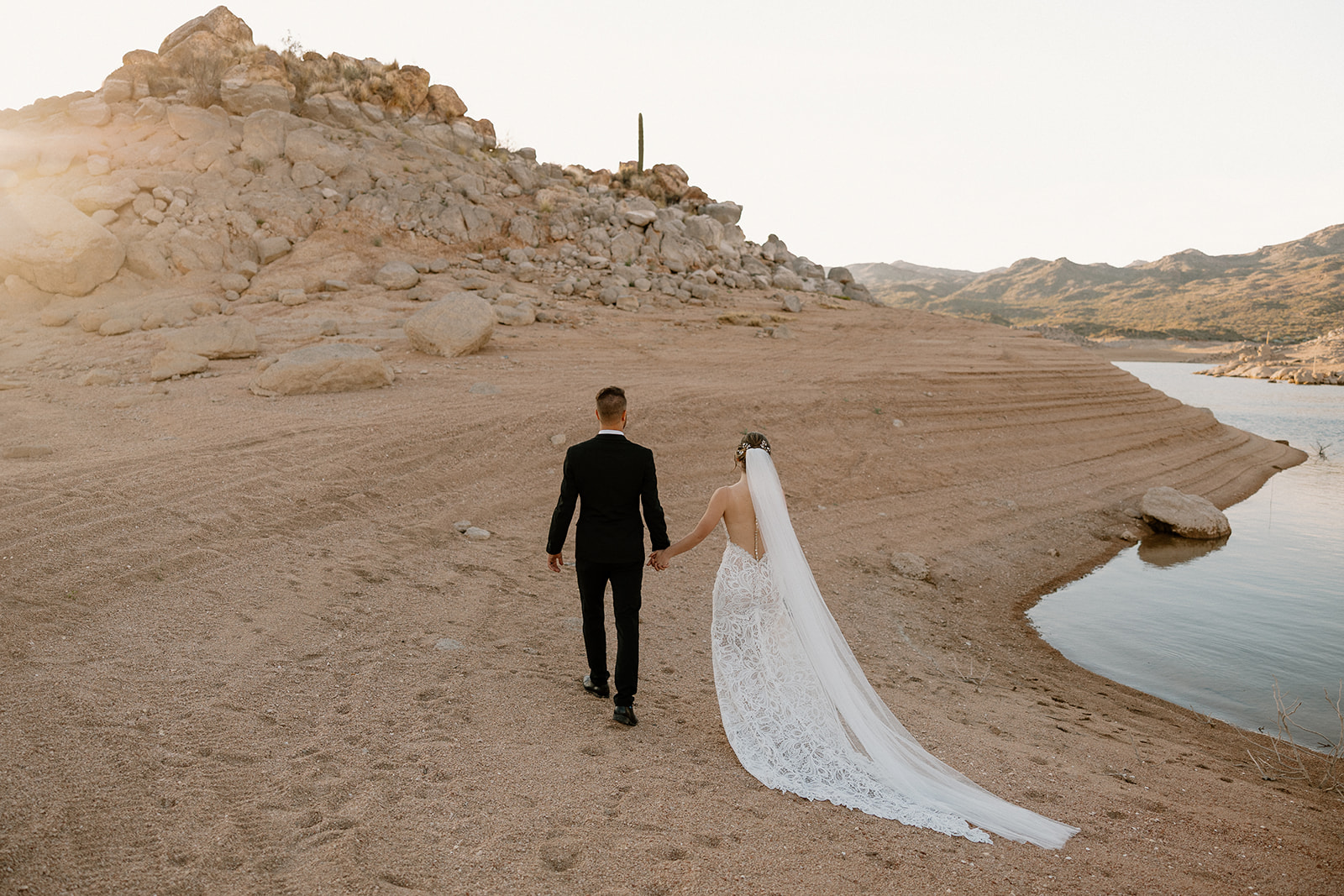Bride and groom walk hand-in-hand along the rocky lakeshore, her long veil trailing behind during their The Cove at Bartlett Lake wedding.
