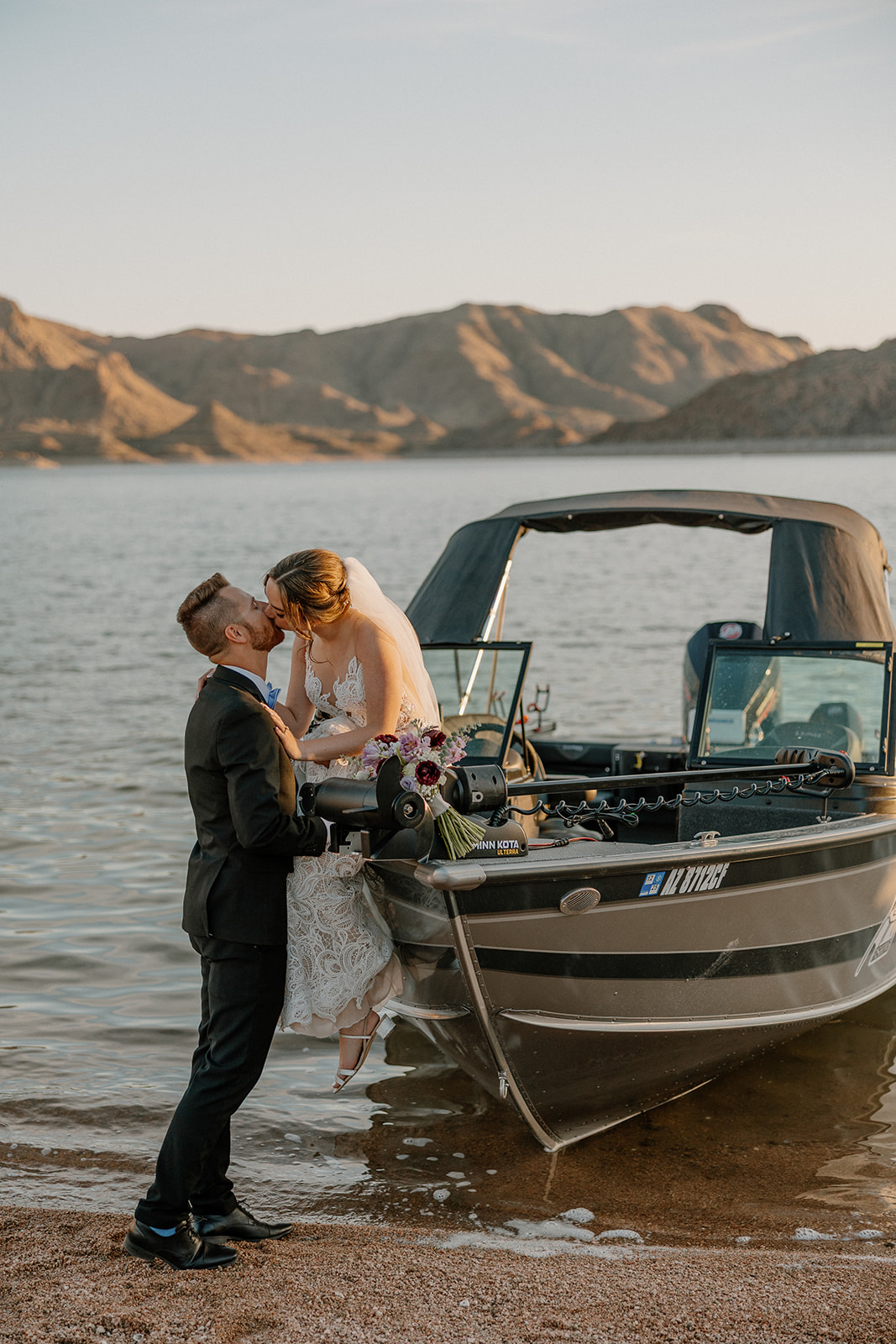 Bride sits on the edge of a boat while the groom kisses her, a romantic lake moment after their wedding ceremony.