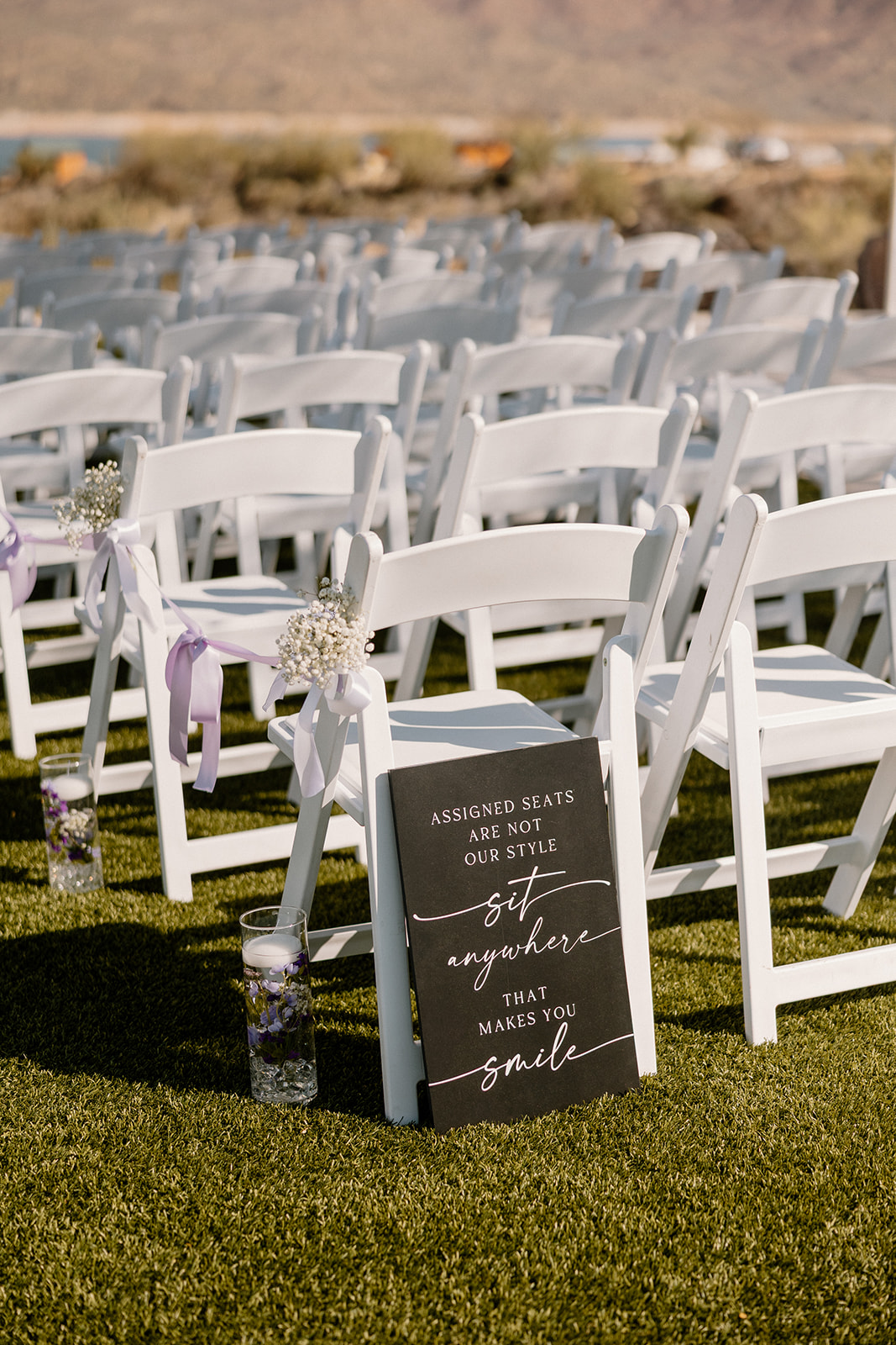 A close-up of white ceremony chairs decorated with baby’s breath and lavender ribbon, with a sign that invites guests to sit together—set against the backdrop of a scenic lake and desert hills at one of the dreamiest waterfront wedding venues in Arizona.