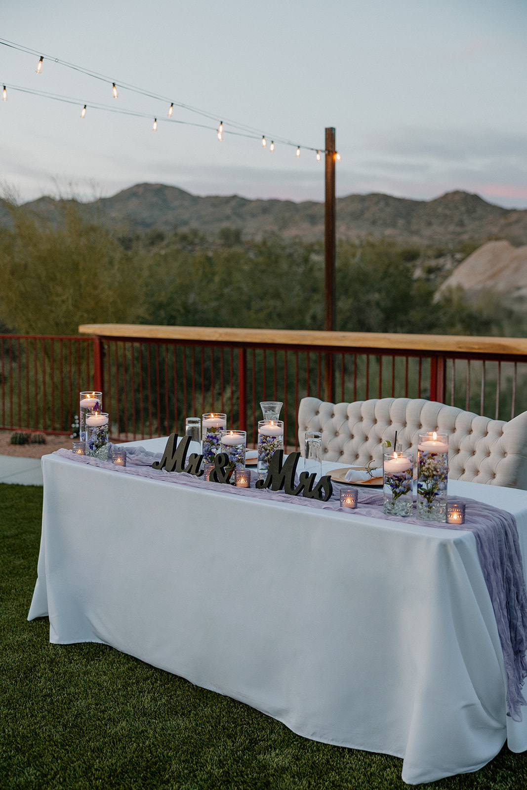A sweetheart table with soft candlelight, lavender accents, and a “Mr. & Mrs.” sign, set outdoors with desert mountain views in the background.