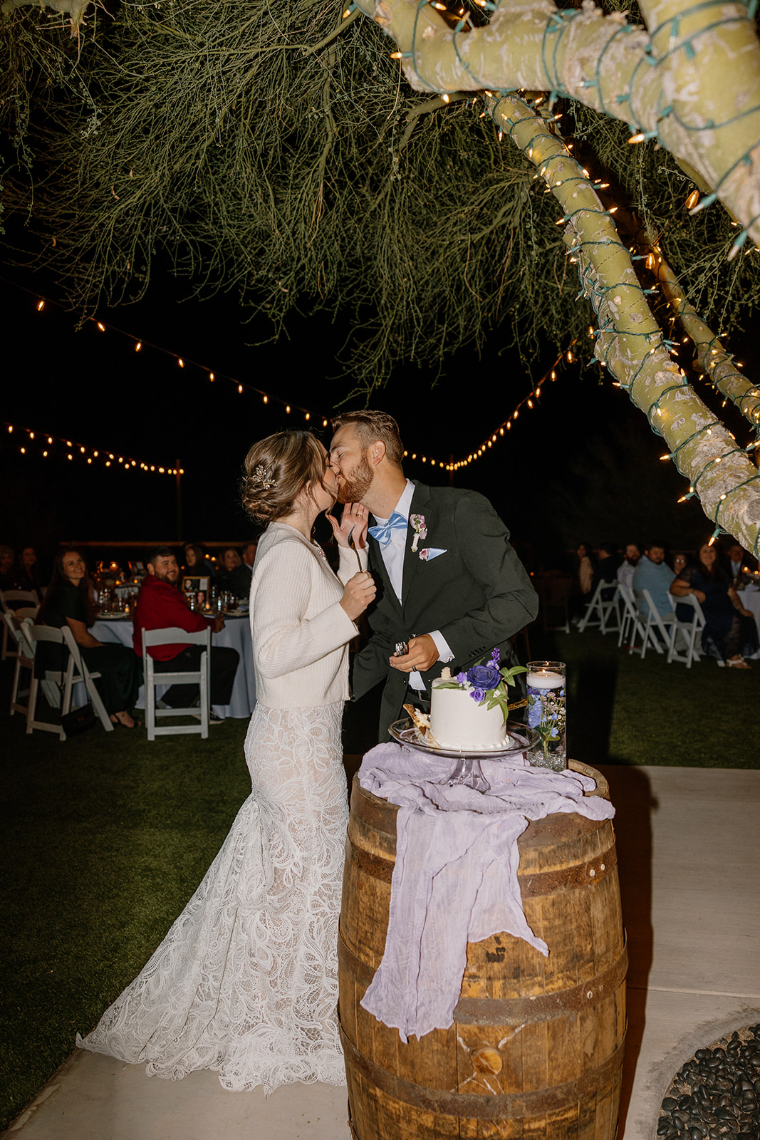 Bride and groom sharing a kiss during their cake cutting under string lights at The Cove at Bartlett Lake wedding reception.

