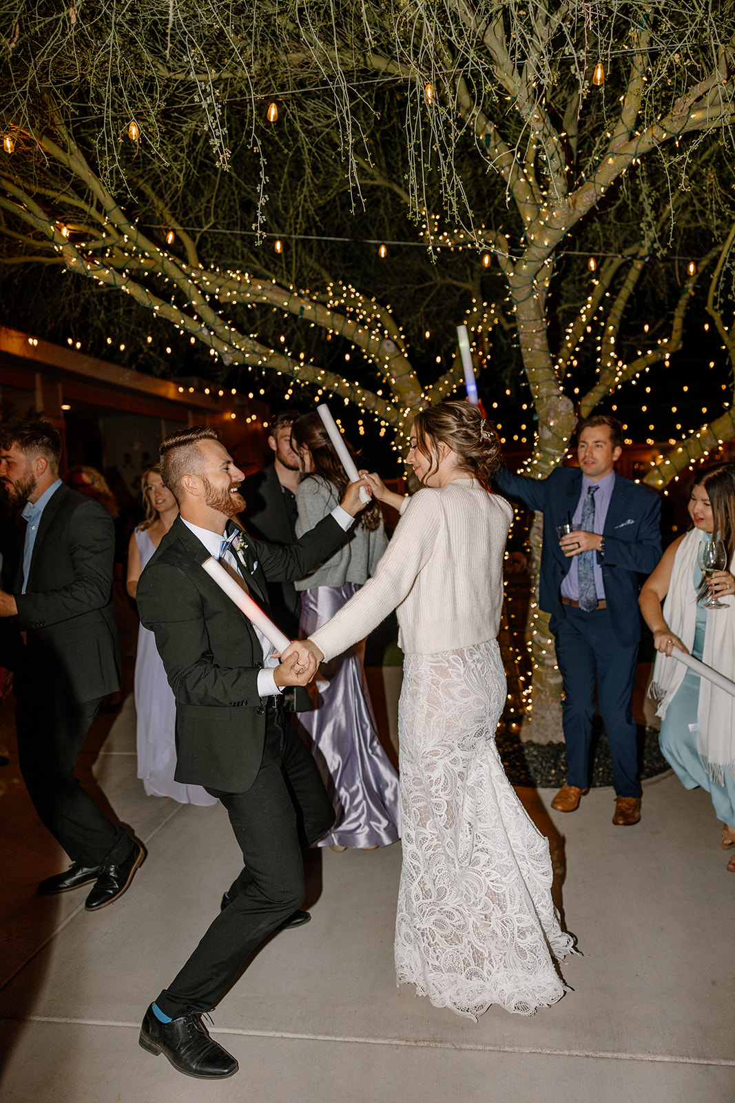 Bride in a lace gown and sweater dancing joyfully with guests under twinkle lights at The Cove at Bartlett Lake wedding.

