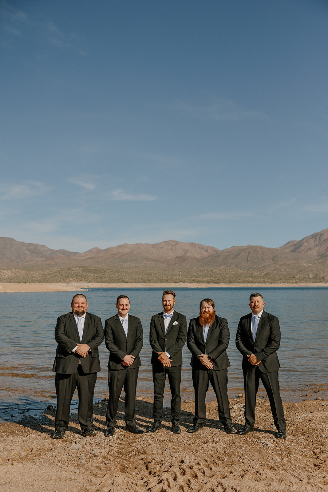 Groom and groomsmen in black suits standing by the shoreline at The Cove at Bartlett Lake wedding with mountain views behind them.

