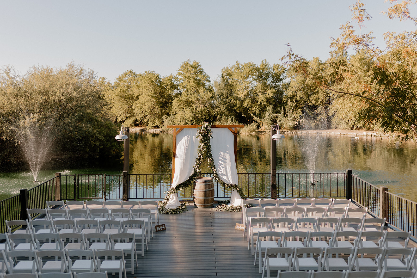 A dreamy ceremony setup with a draped wooden arch, white chairs, and a lake backdrop framed by fountains and lush trees—perfect for couples eyeing waterfront wedding venues in Arizona.
