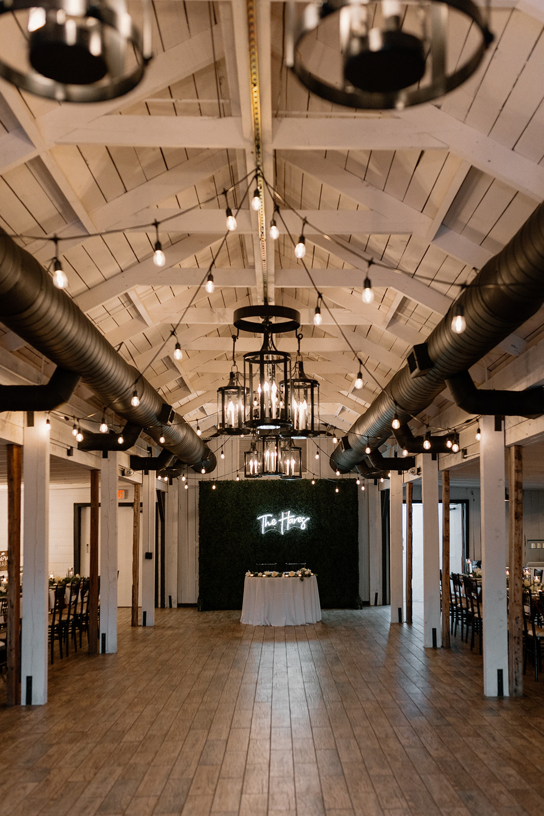 A modern rustic wedding reception space with exposed beams, string lights, and a mossy green wall featuring a neon sign—elegant and cozy indoor setup.