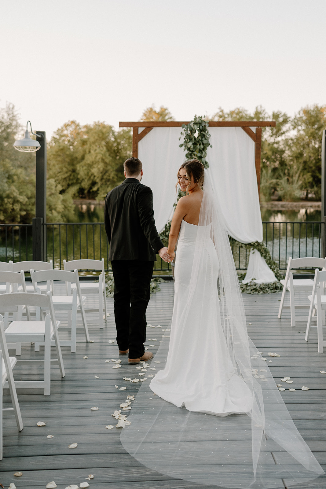 A bride and groom hold hands and smile as they walk down the aisle toward a wooden altar by the water—one of the most peaceful waterfront wedding venues in Arizona.
