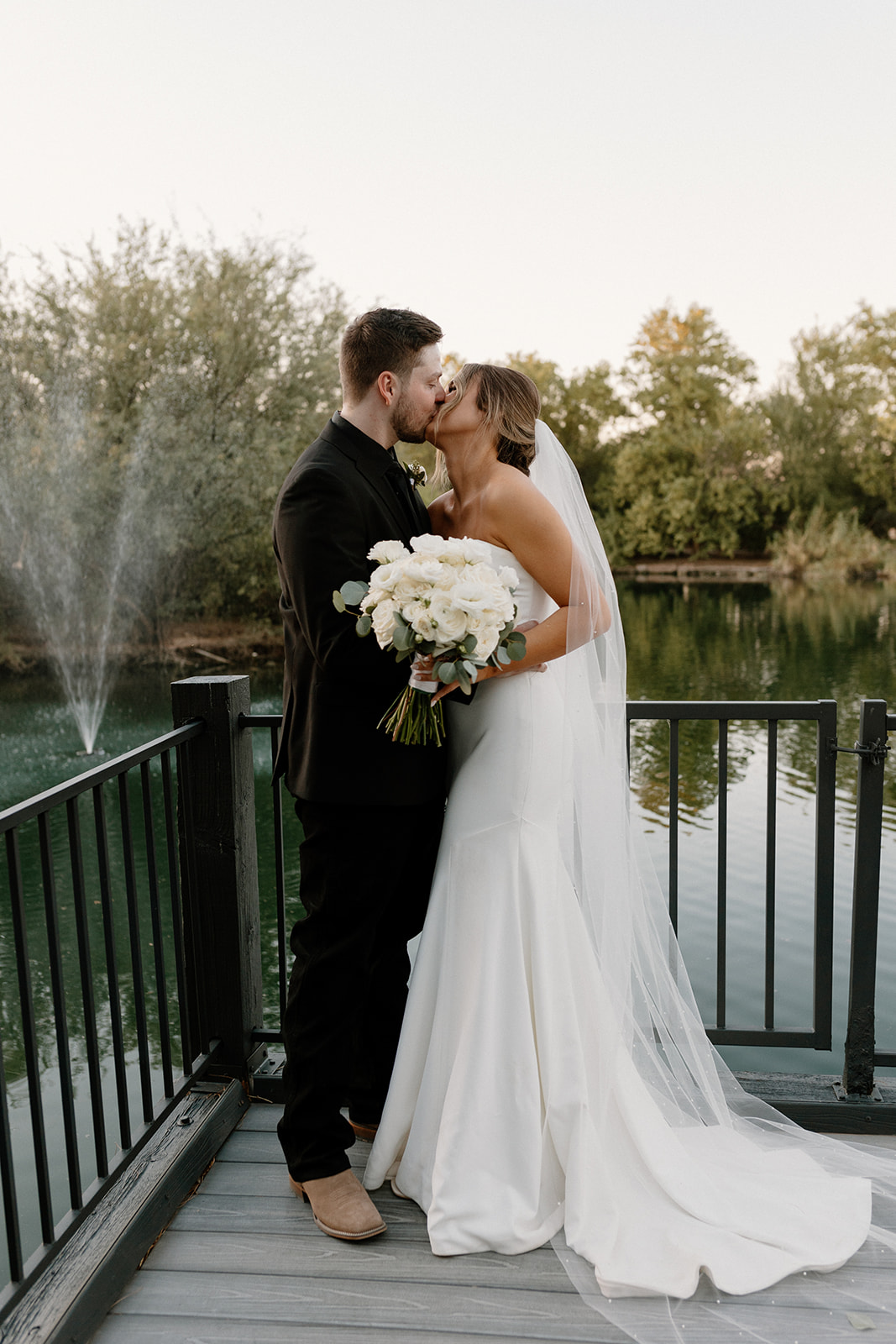 The bride and groom share a kiss on a dock surrounded by still water and a nearby fountain—serene beauty at its finest from waterfront wedding venues in Arizona.