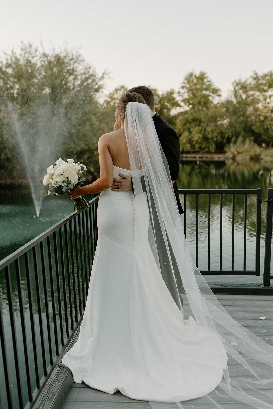 A bride and groom embrace on a dock with a fountain spraying in the background, showing off the peaceful beauty of waterfront wedding venues in Arizona.