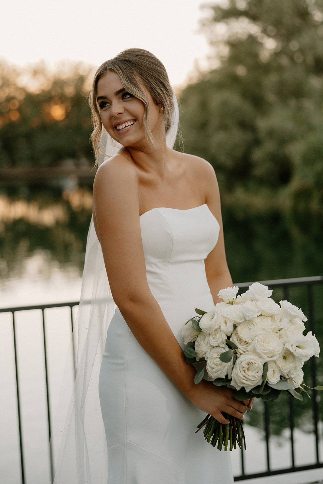 A smiling bride in a strapless white gown holding a bouquet of white roses, standing in front of a serene lake surrounded by lush greenery.