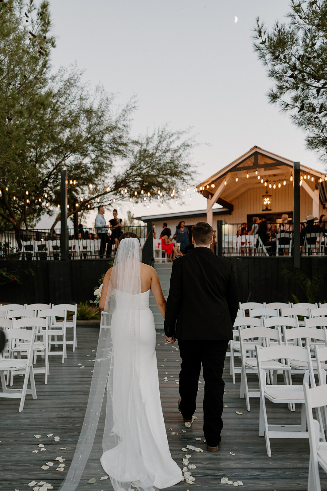 A couple walks down a white-petal-strewn aisle at an outdoor ceremony with string lights and a live band in the background—romantic vibes at a lakeside venue