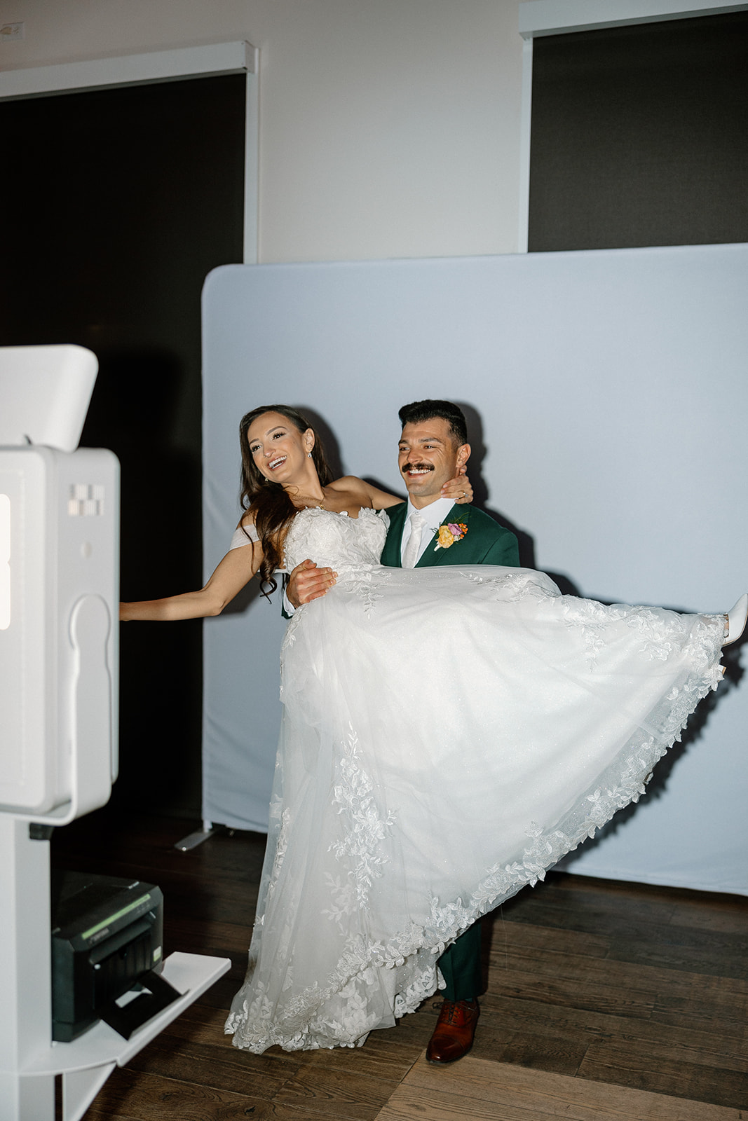 The groom lifts the bride in front of a photo booth backdrop as they laugh together during the reception.

