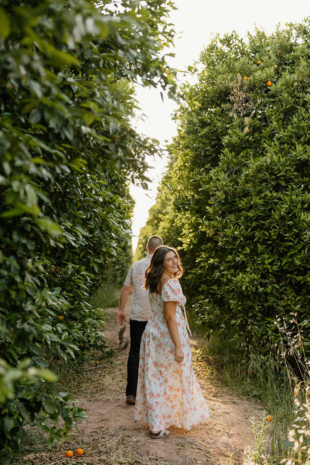 Couple walking through green orchard of oranges while woman looks back smiling