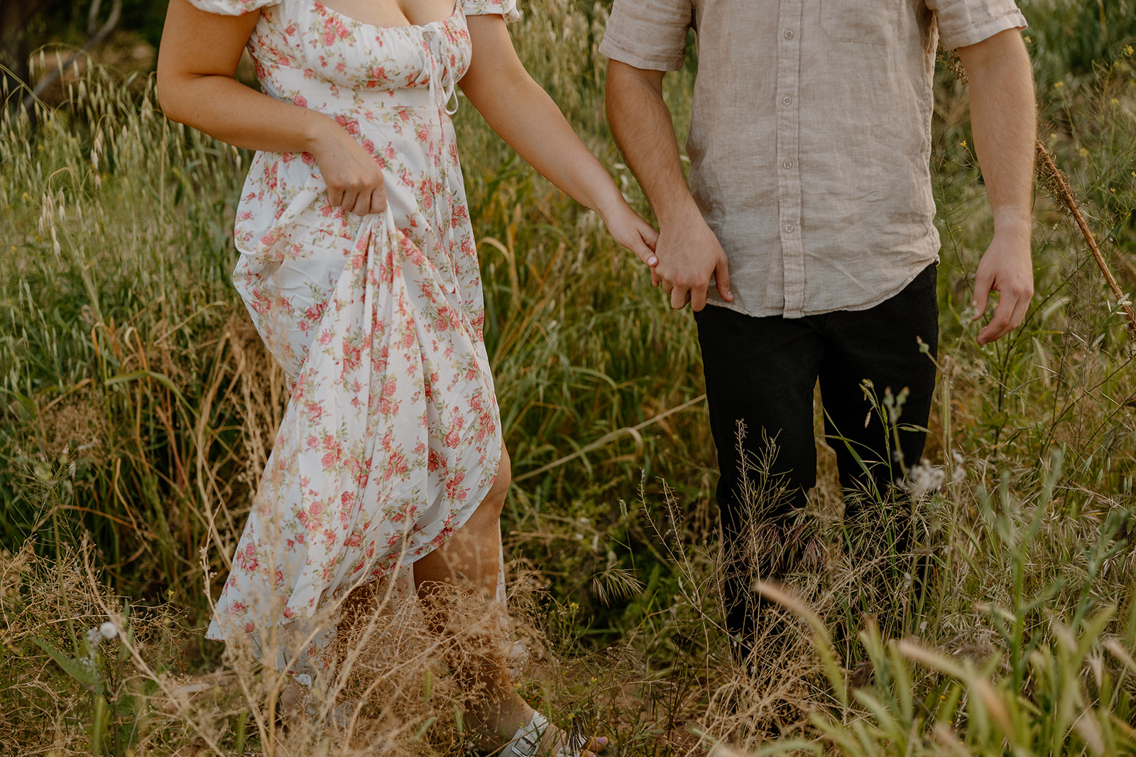 Newly engaged couple walking through tall green grass hand in hand while woman holds her dress