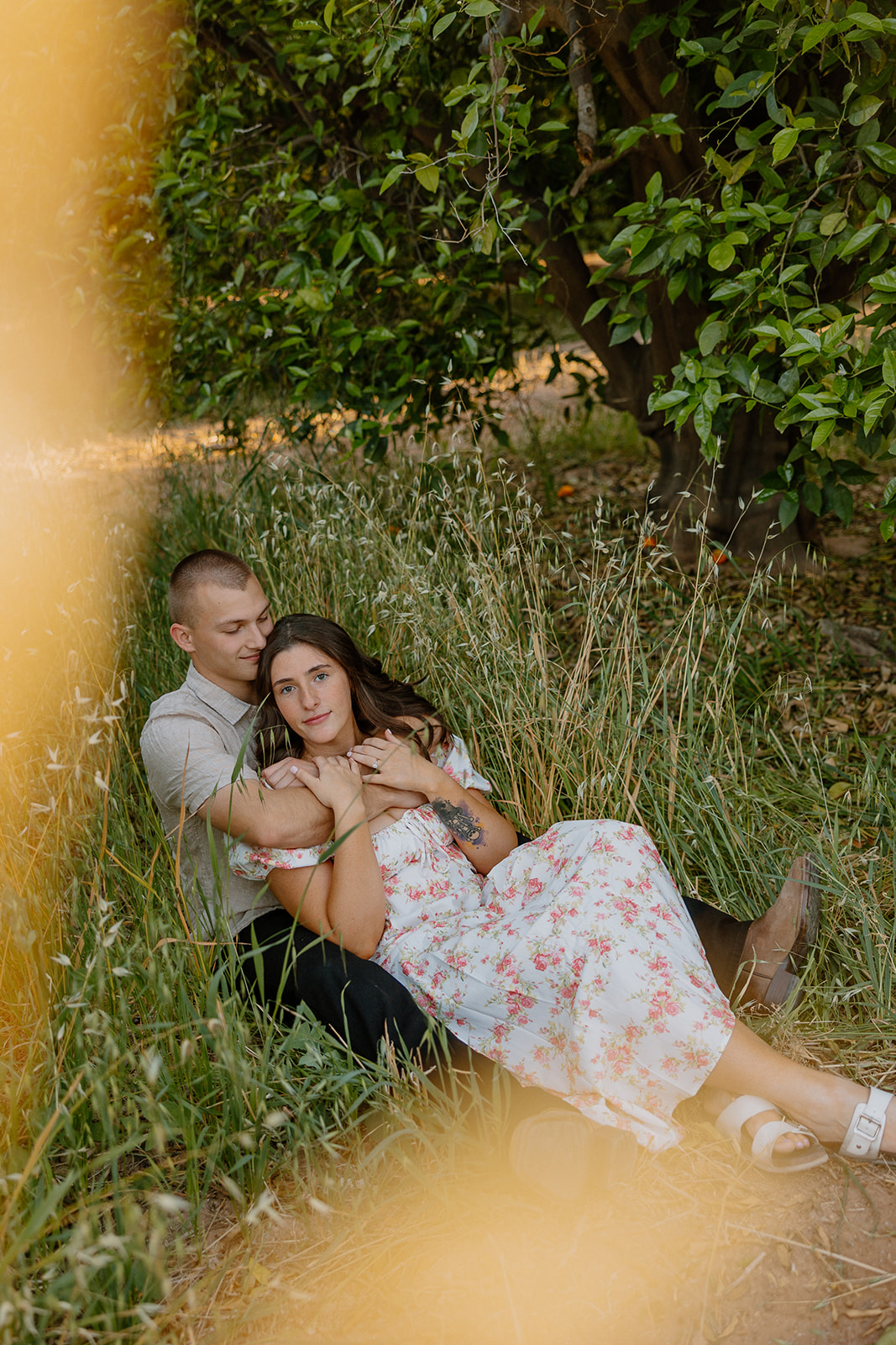 Couple sitting in green grass outside orchard during engagement photos
