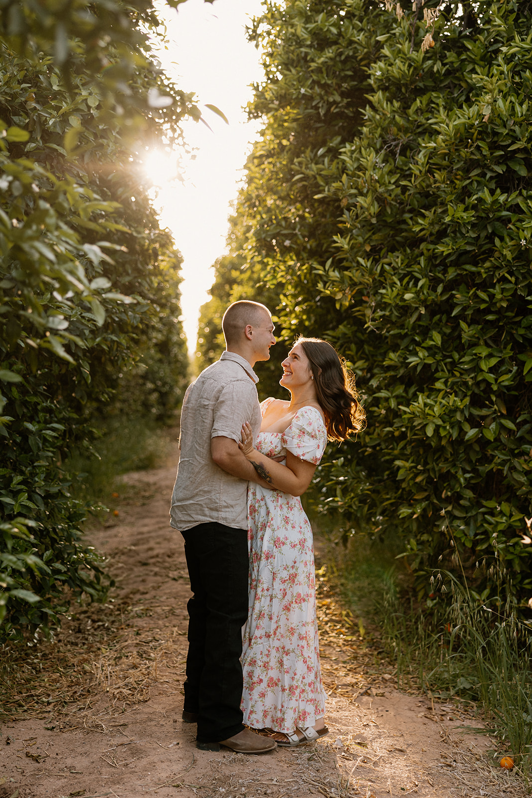 Golden hour orchard engagement photos with woman wearing white floral dress and man wearing casual outfit