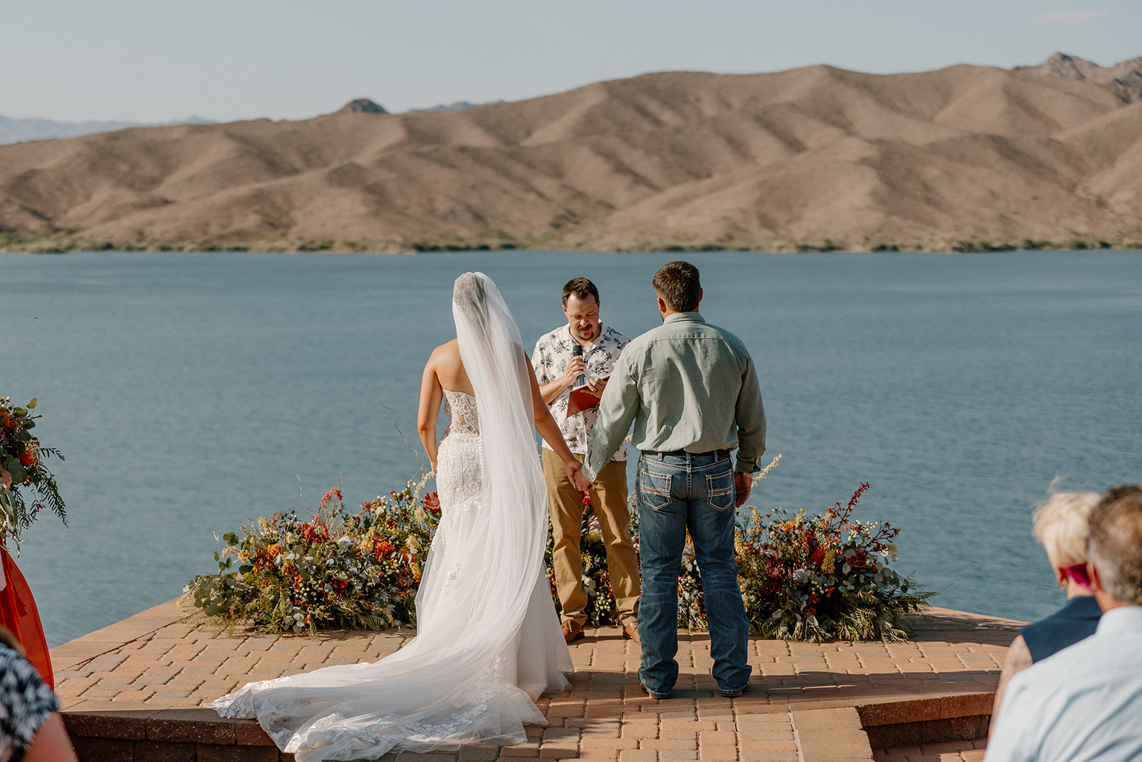 A couple exchanges vows on a brick platform framed by colorful florals, with a vast lake and rugged Arizona mountains behind them—an iconic scene from a waterfront wedding venue in Arizona.