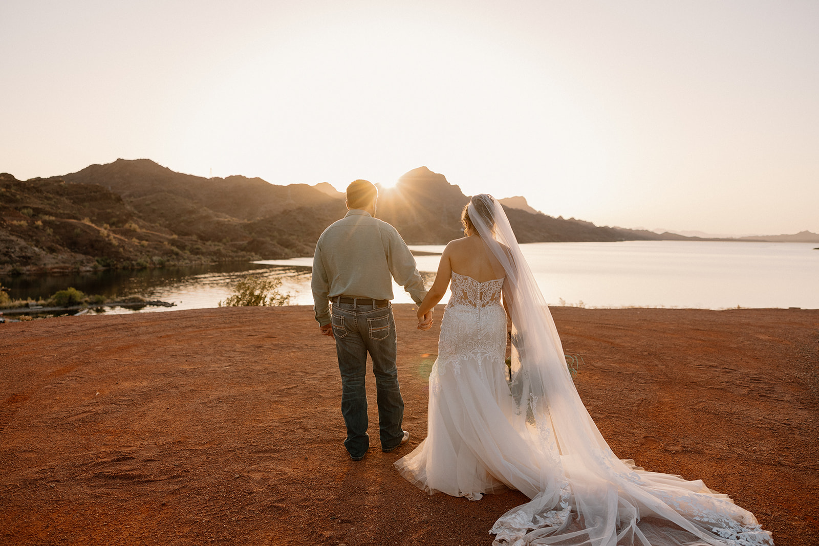 A bride in a lace gown and a groom in jeans and a button-up shirt walk hand-in-hand toward the water at sunset, capturing the charm of waterfront wedding venues in Arizona.