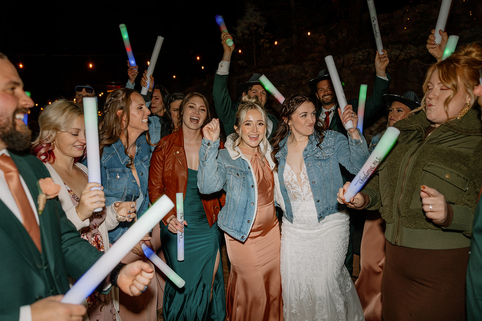 Guests in glow gear and denim jackets dance with the bride in the center of a high-energy outdoor wedding party.

