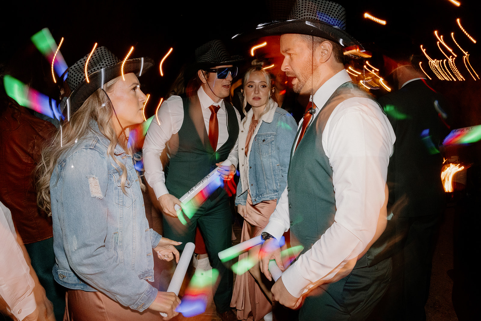 Wedding party guests in cowboy hats and glow sticks dance under neon lights at an Arizona wedding reception.


