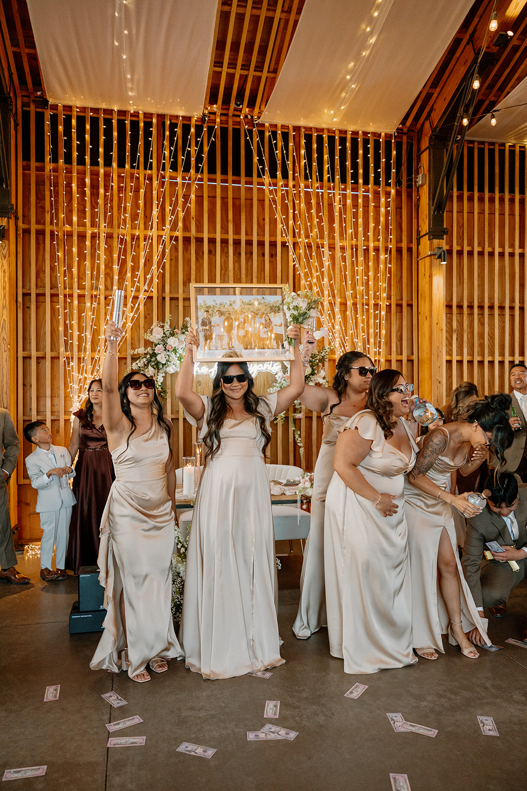 Bridesmaids in champagne satin dresses celebrate with glow sticks and a framed photo in front of dreamy string lights.

