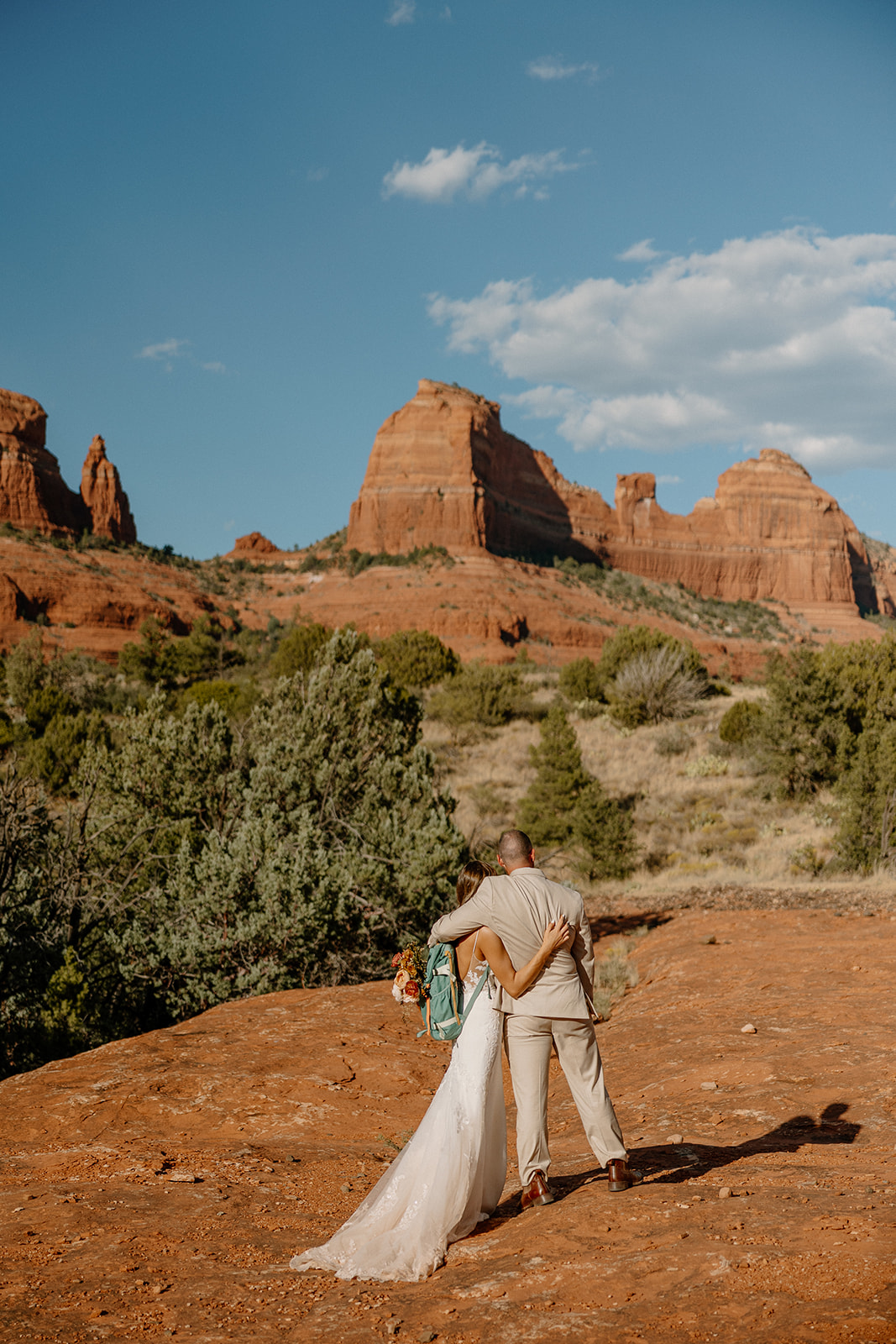 Bride and groom stand hand in hand, gazing out over red rock spires during their arizona adventure elopement in the desert sun.